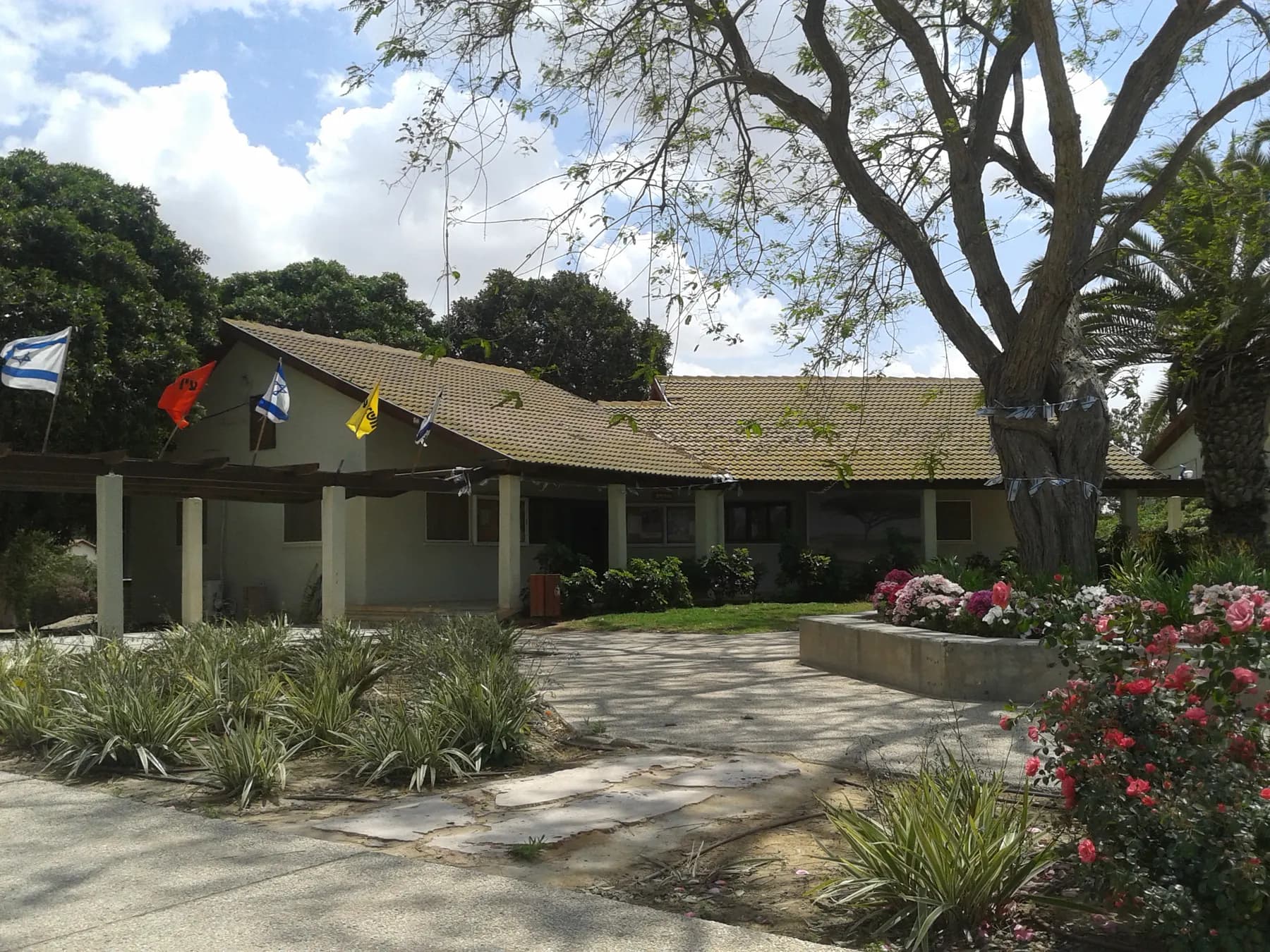 A low building with a tiled roof and flags over its porch in Ein HaBesor, behind a garden of shrubs and flowering roses.
