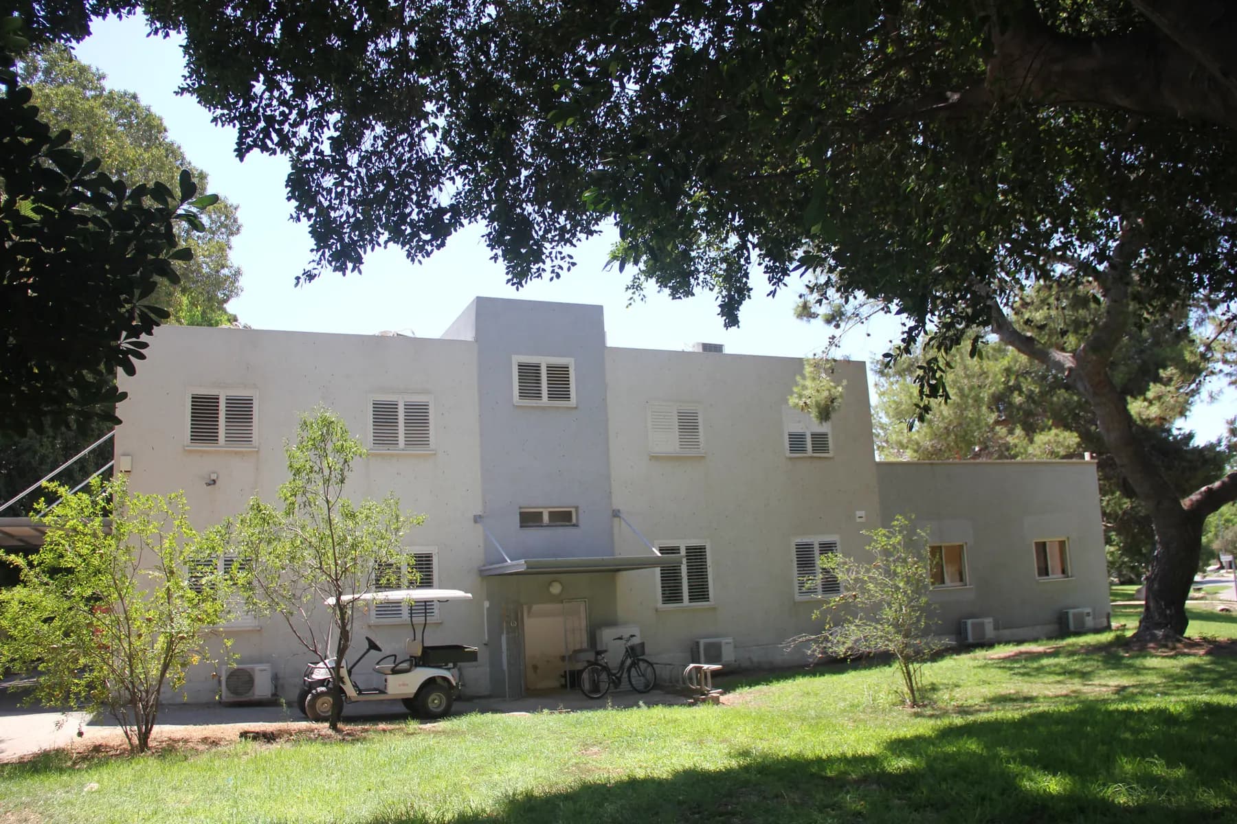 A white two-storey building on a lawn in Ein HaMifratz, shaded by a large tree, with a golf cart and bicycles parked by the entrance.