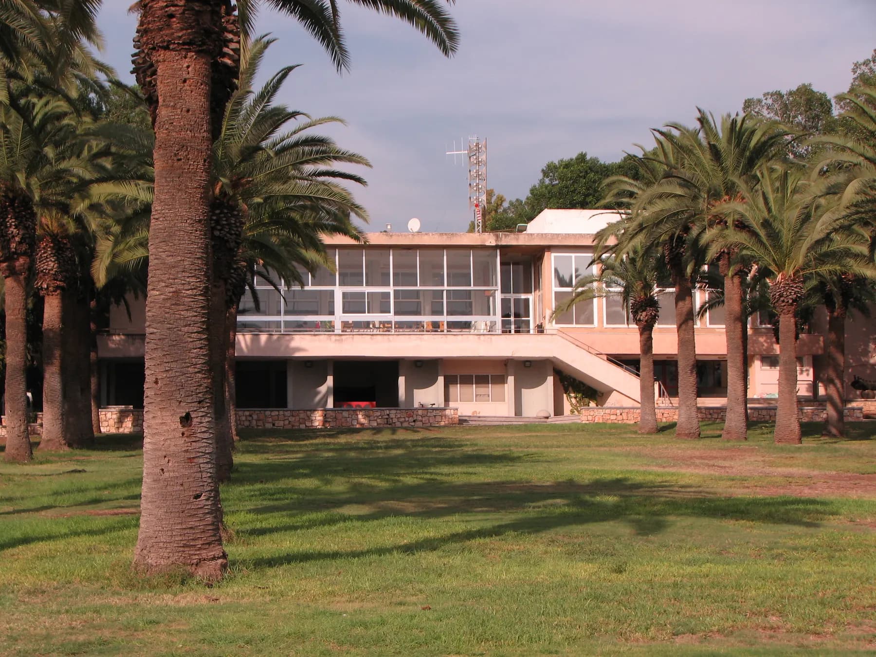 The kibbutz dining hall in Ein Harod Meuhad, a long building with wide windows, behind a lawn with tall palm trees.