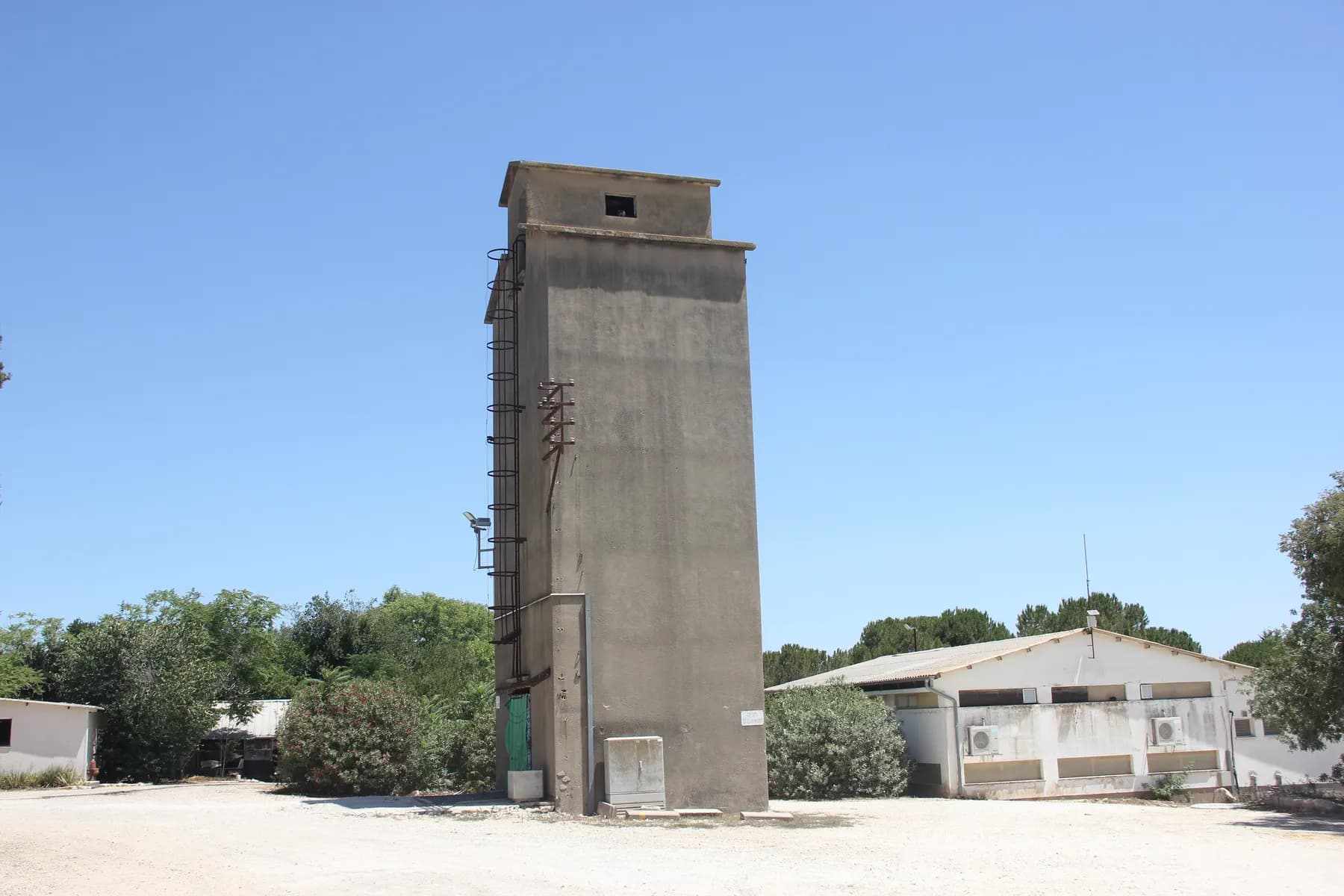 A tall concrete tower with a ladder up its side beside a low farm building in Ein HaShofet, under a clear sky.