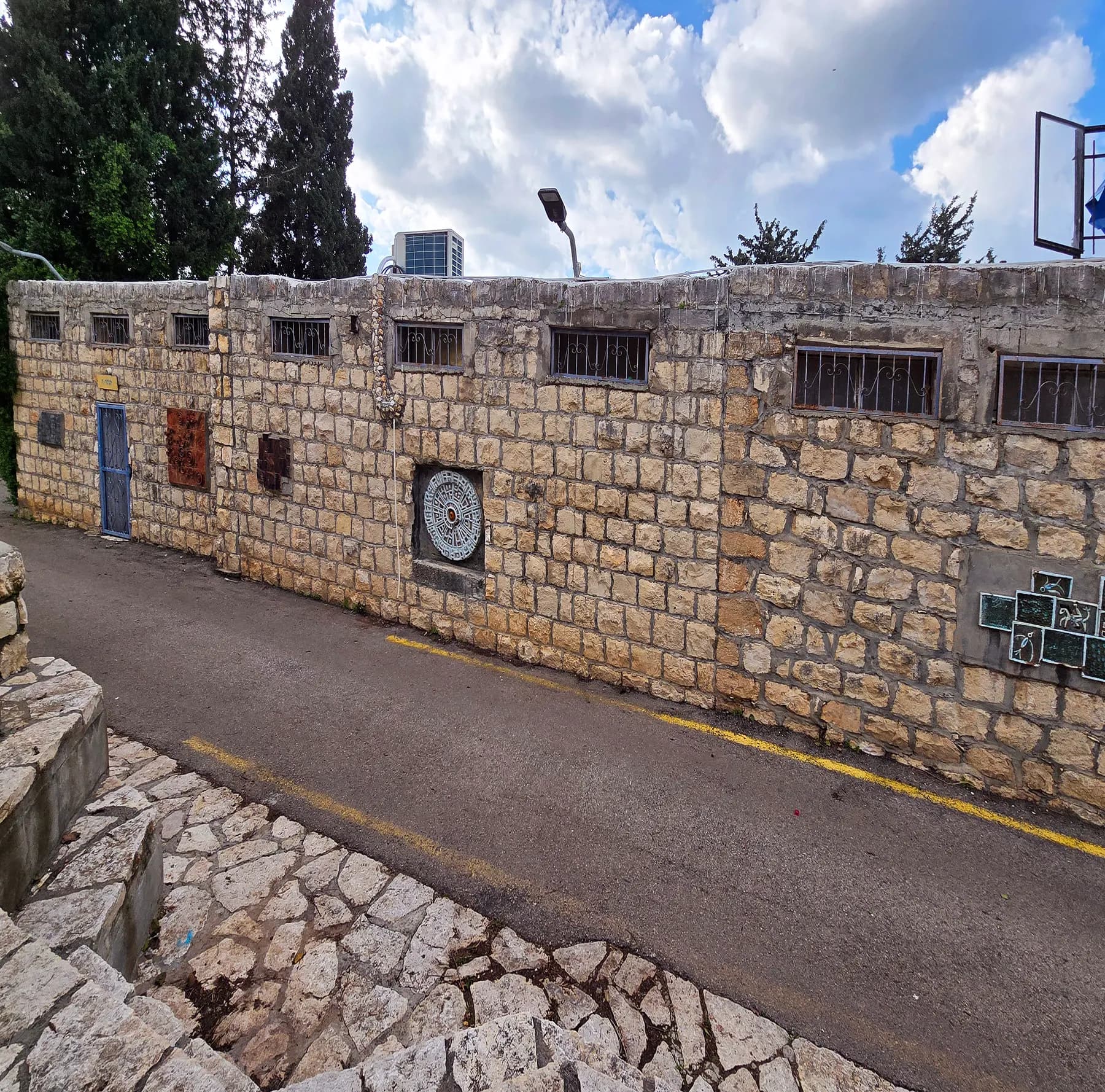 A long stone building with barred windows and artworks mounted on its wall beside a lane in Ein Hod.