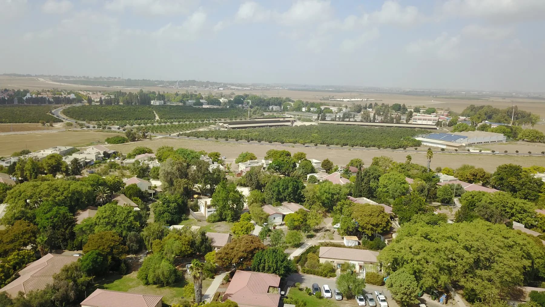 An aerial view over Ein Tzurim, small red-roofed houses among dense trees, with orchards, fields and farm buildings stretching to a hazy horizon.