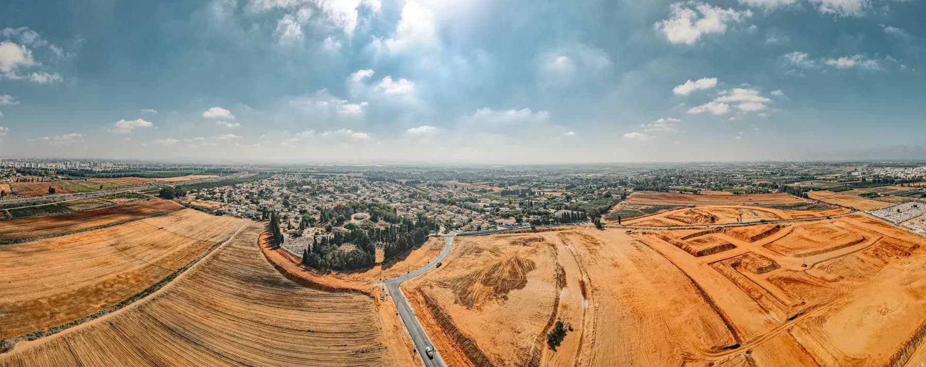 A drone view over the houses and trees of Elyakhin, with bare earthworks for new building and a road in the foreground.