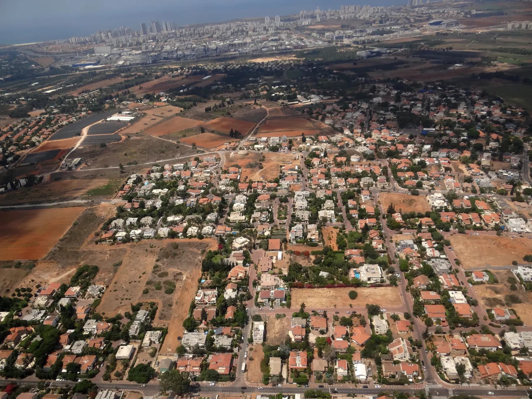 An aerial view of red-roofed houses and tree-lined streets in Even Yehuda, with fields and distant city towers on the horizon.