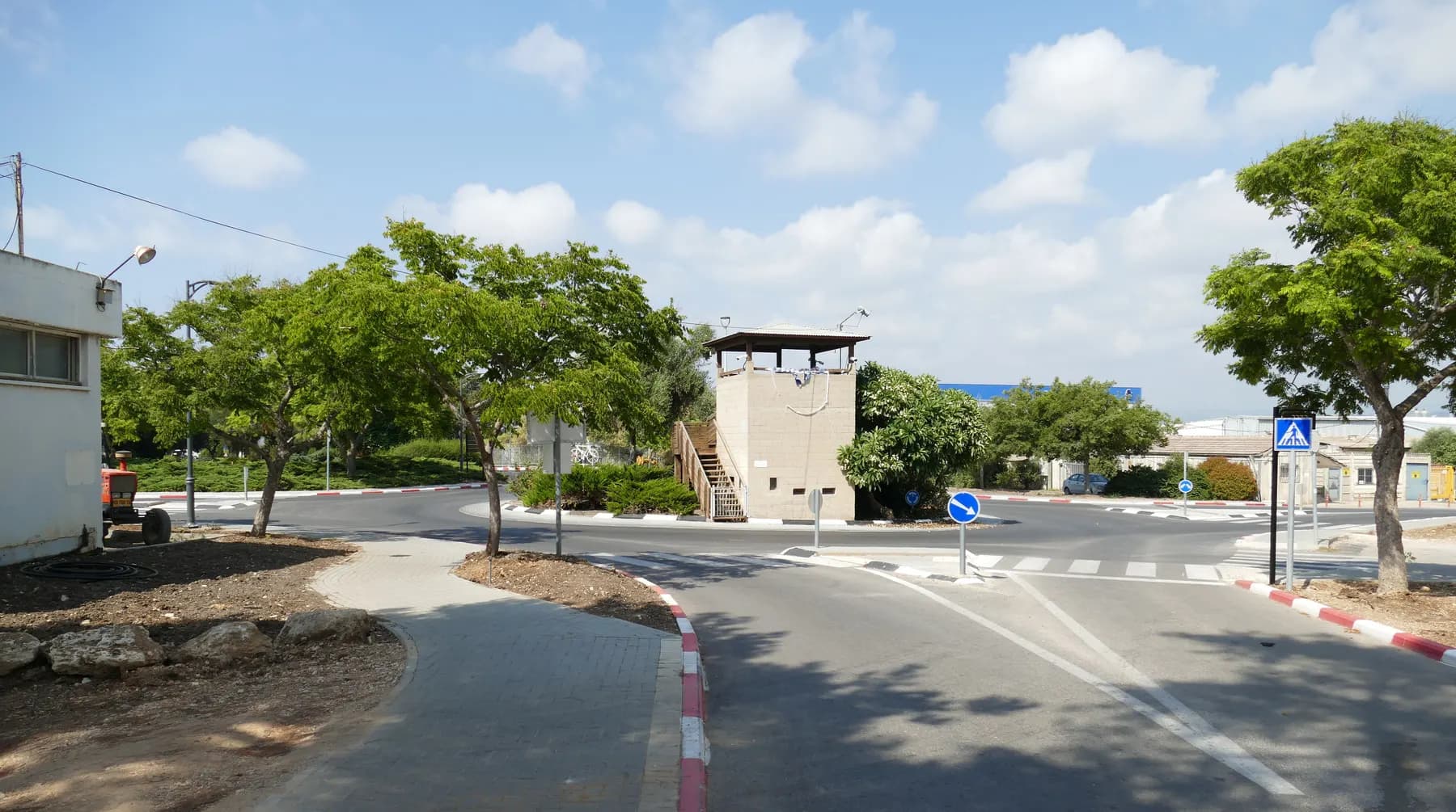 A road junction in Evron with a square watchtower, reached by an outdoor staircase and topped with a roofed lookout, among leafy trees.