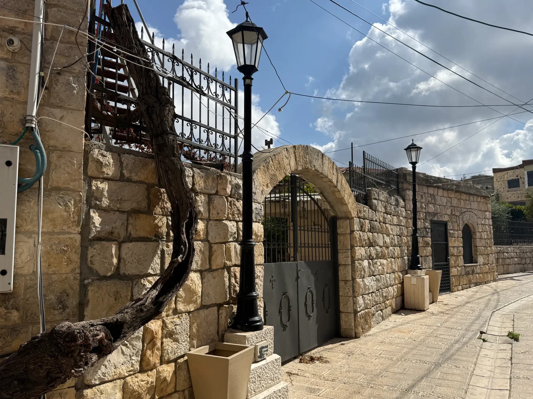 A quiet lane in Fassuta between walls of pale stone, with an arched iron gate, black street lamps and an old twisted tree trunk.