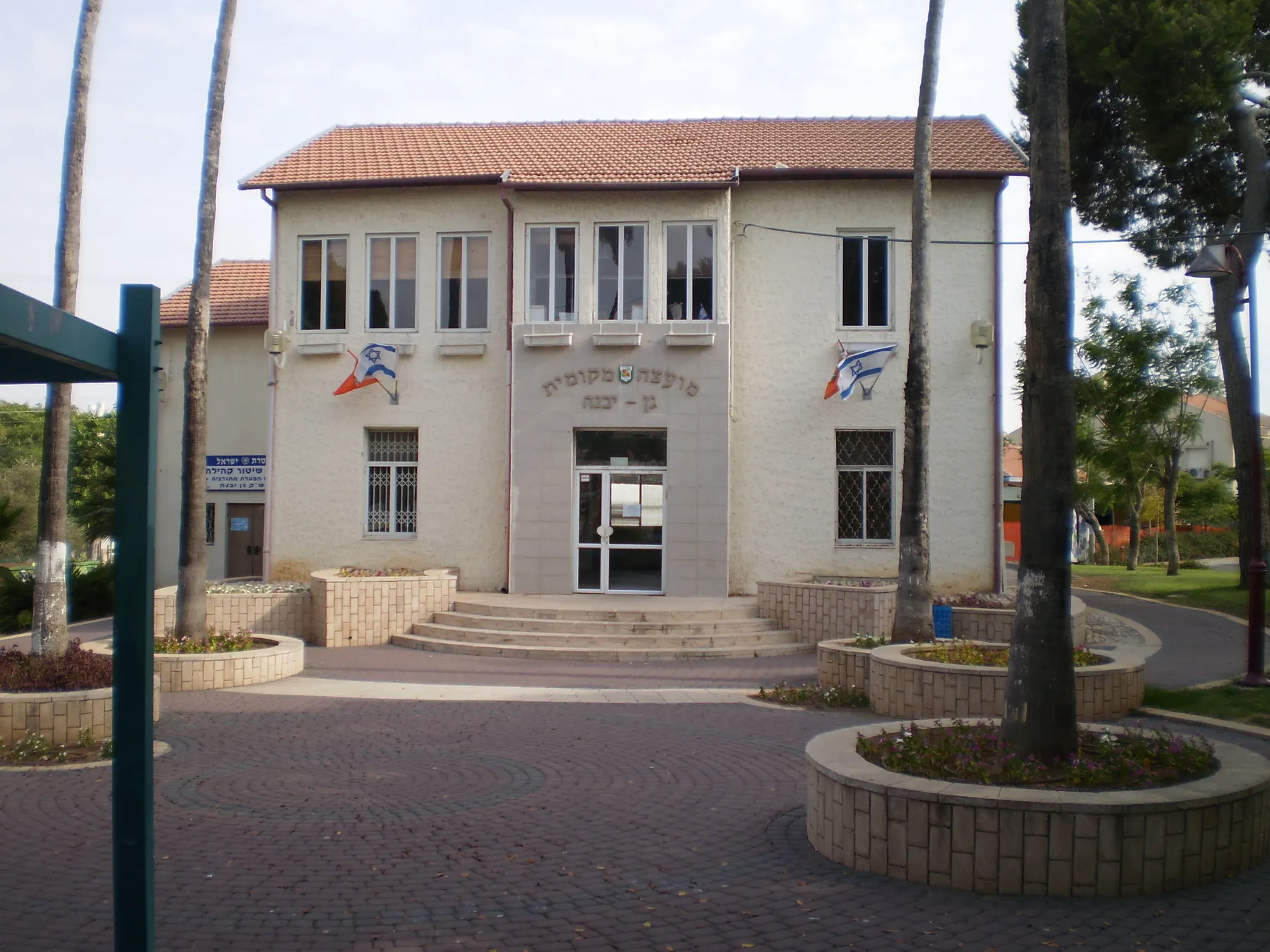 A two-storey building with a red tiled roof facing a paved square with palm trees in Gan Yavne.