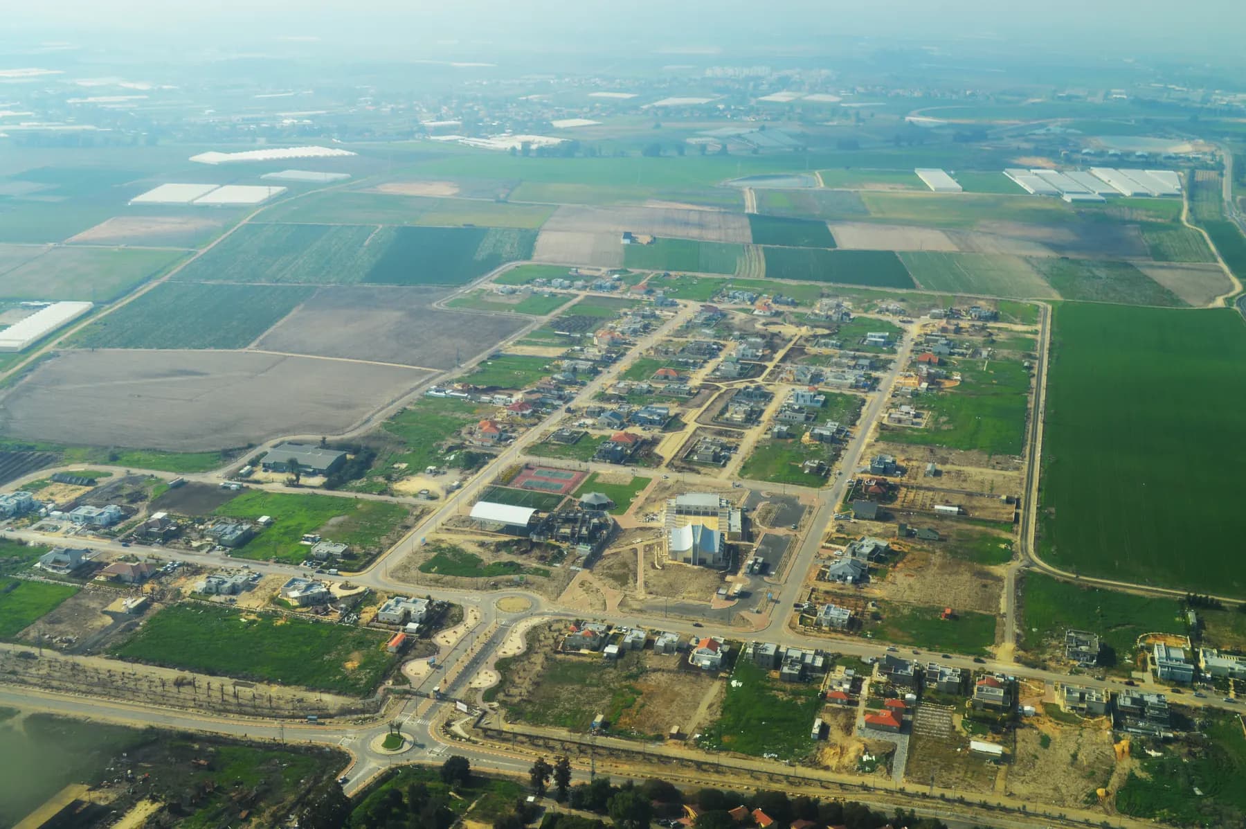 An aerial view of Ganei Tal, with new houses and empty plots along a grid of streets and green fields all around.