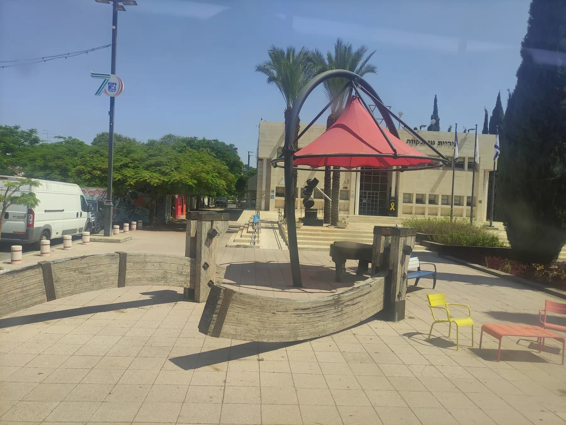 The paved plaza in front of City Hall in Ganei Tikva, with a red shade canopy, palm trees and benches.