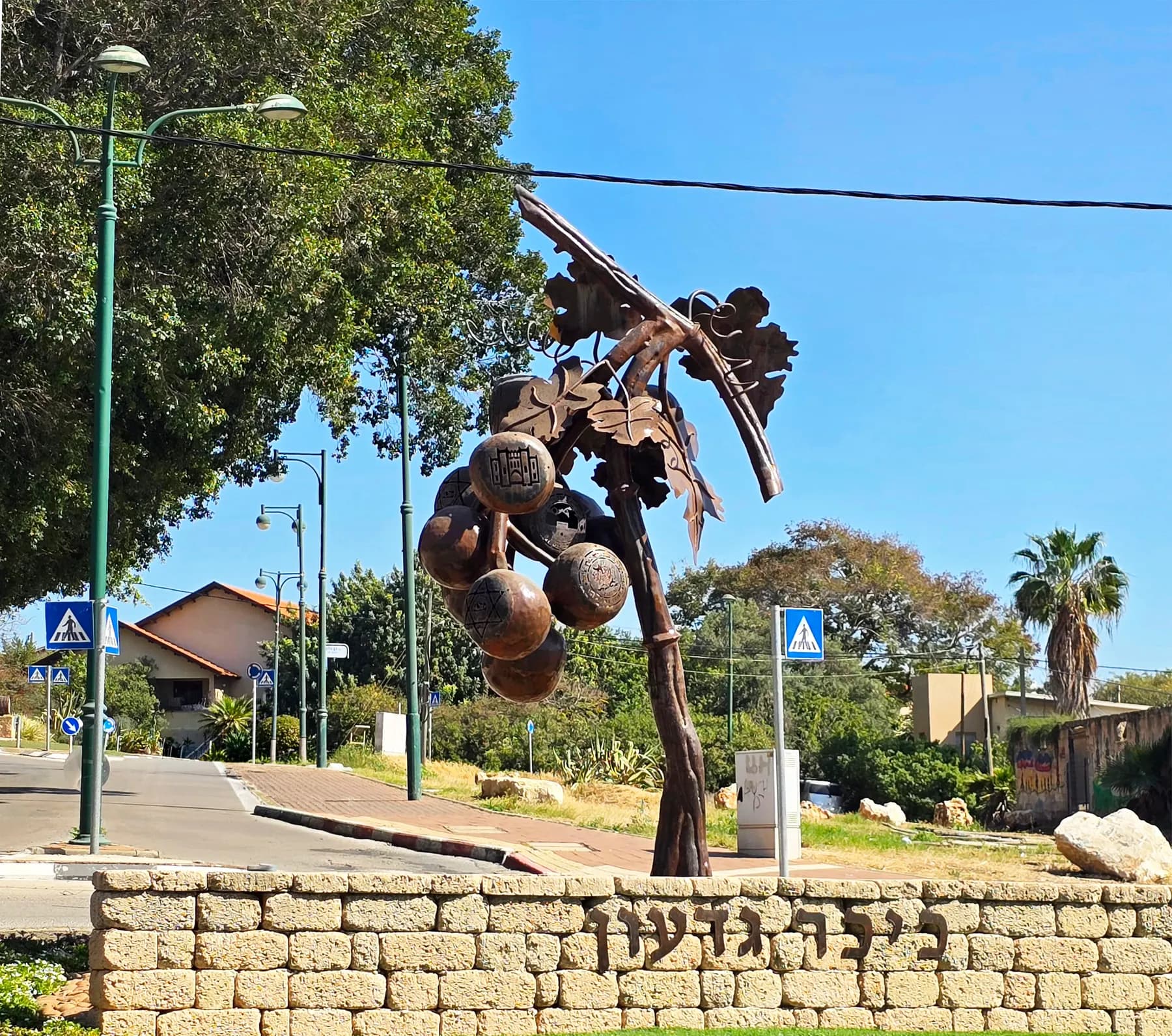 A tall metal sculpture of a vine hung with large round fruit on Gideon Square in Gedera, with palms and red-roofed houses behind.
