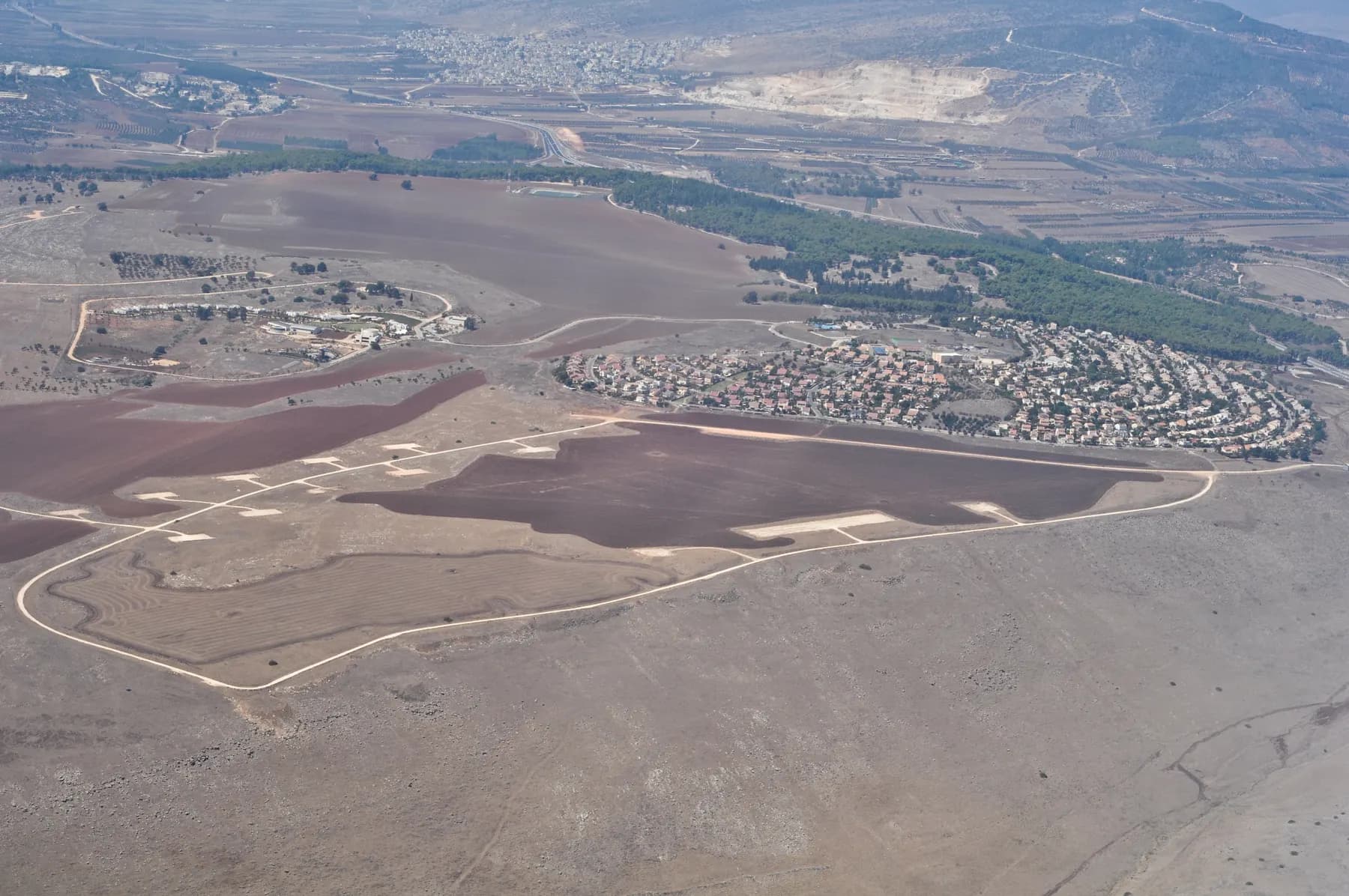 An aerial view of Giv'at Avni, its houses laid out along curving streets beside wide ploughed fields, with a belt of pine trees and hills with a quarry beyond.