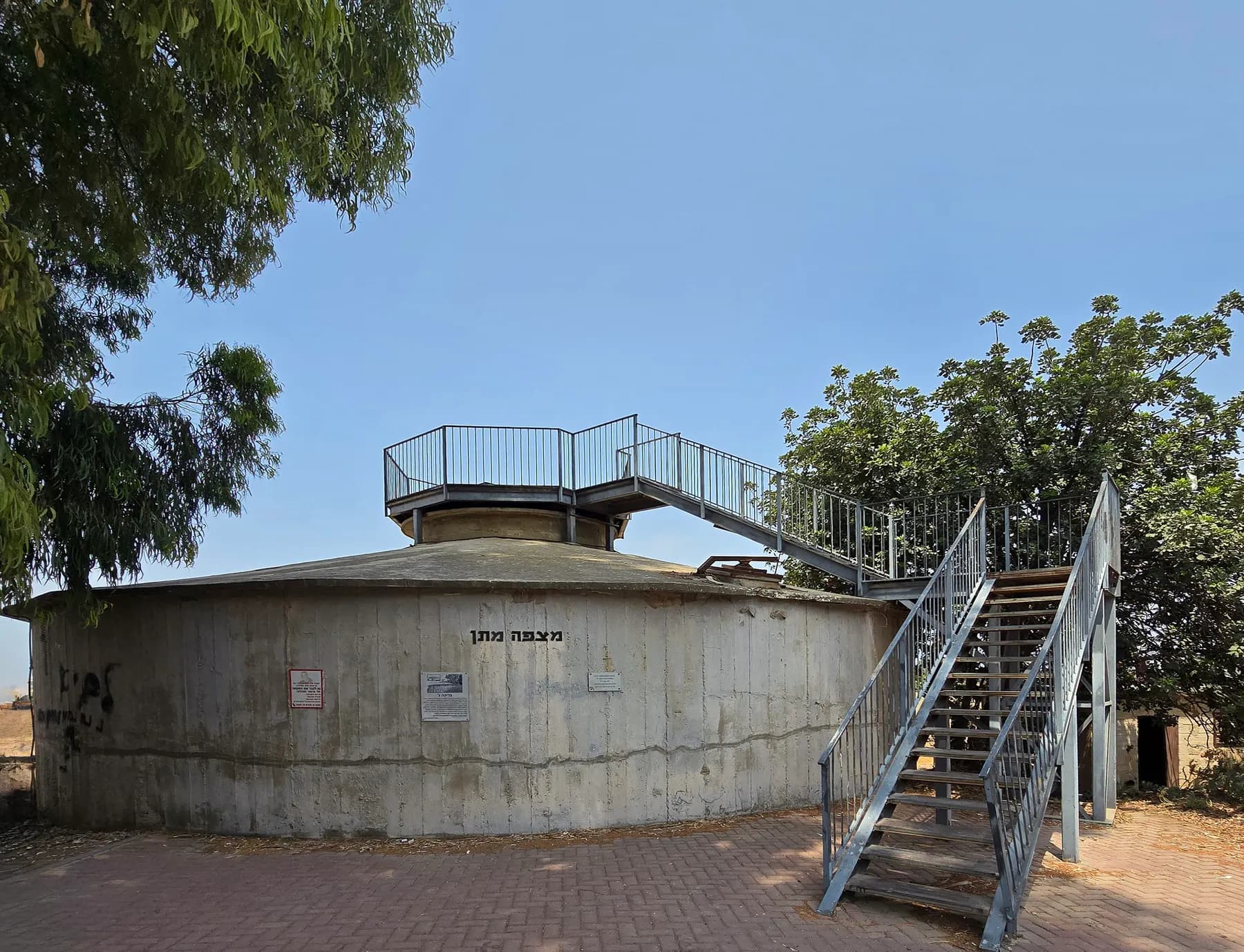A low round concrete water pool at the edge of Givat Brenner, with a metal staircase climbing to a railed lookout on its roof and trees around it.