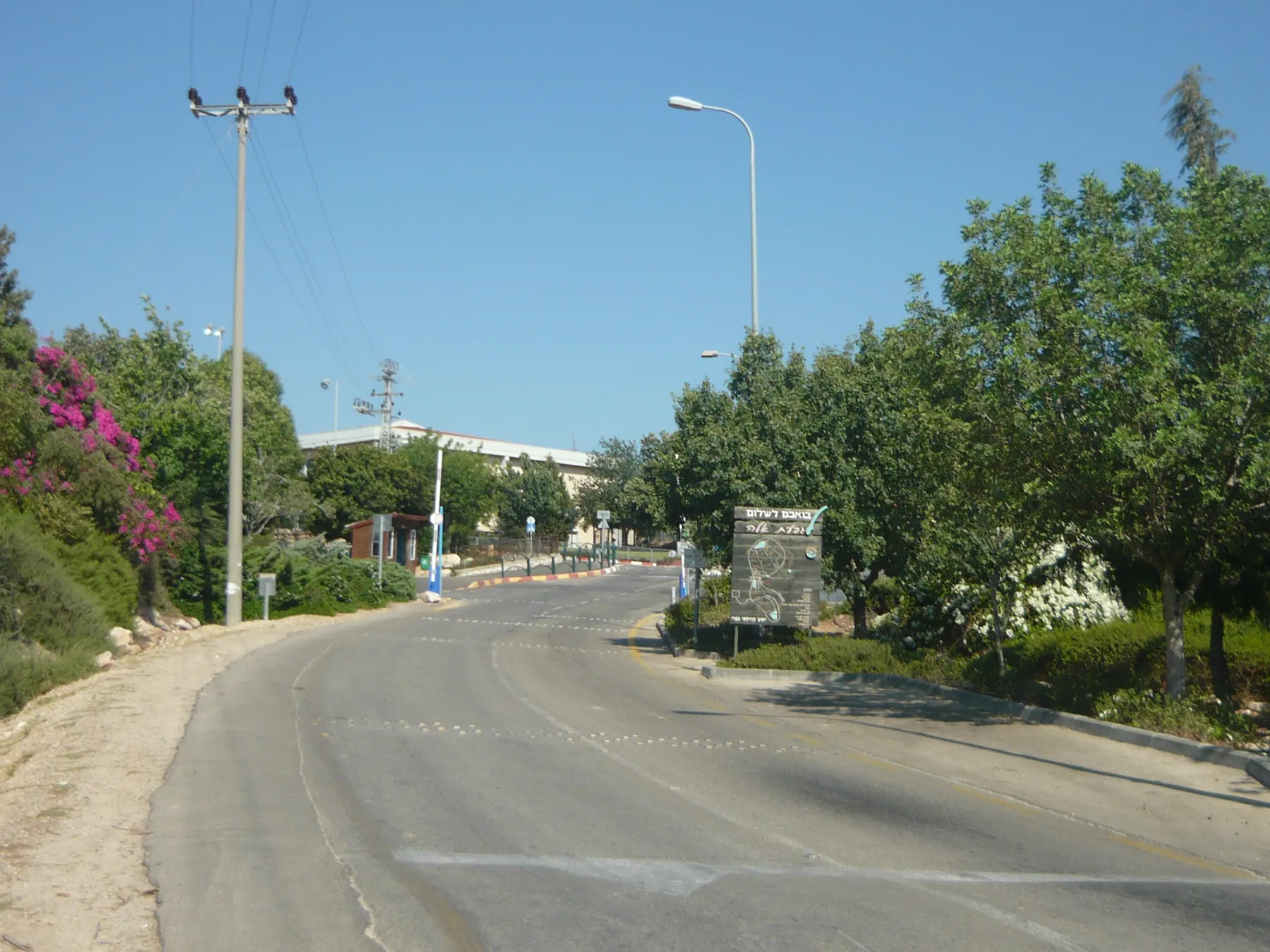 The entrance road into Givat Ela, curving uphill between leafy trees and a pink bougainvillea, with a sign by the roadside and low buildings ahead.