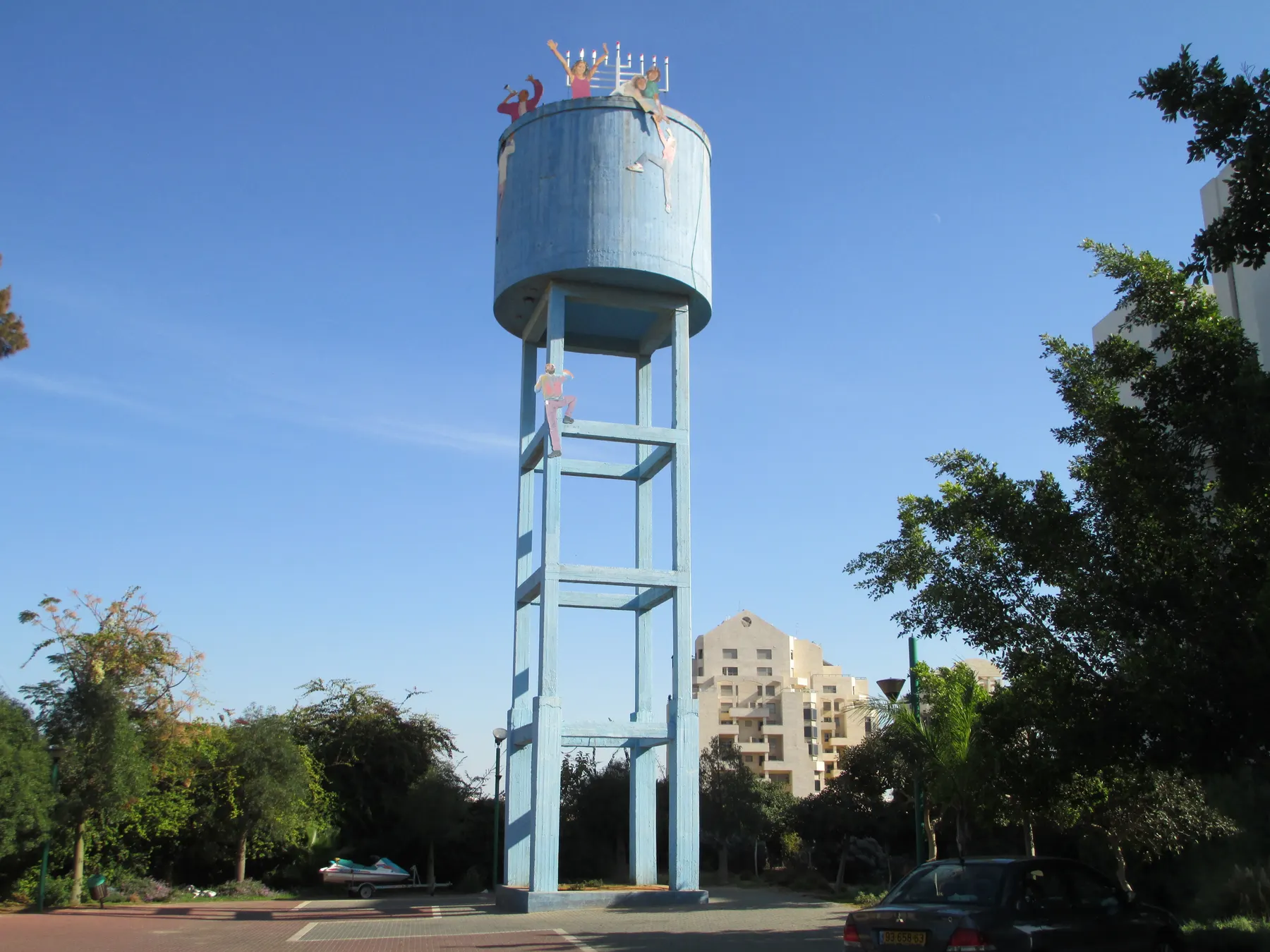 The old blue water tower of Givat Shmuel with colourful figures on it, among trees and apartment buildings.