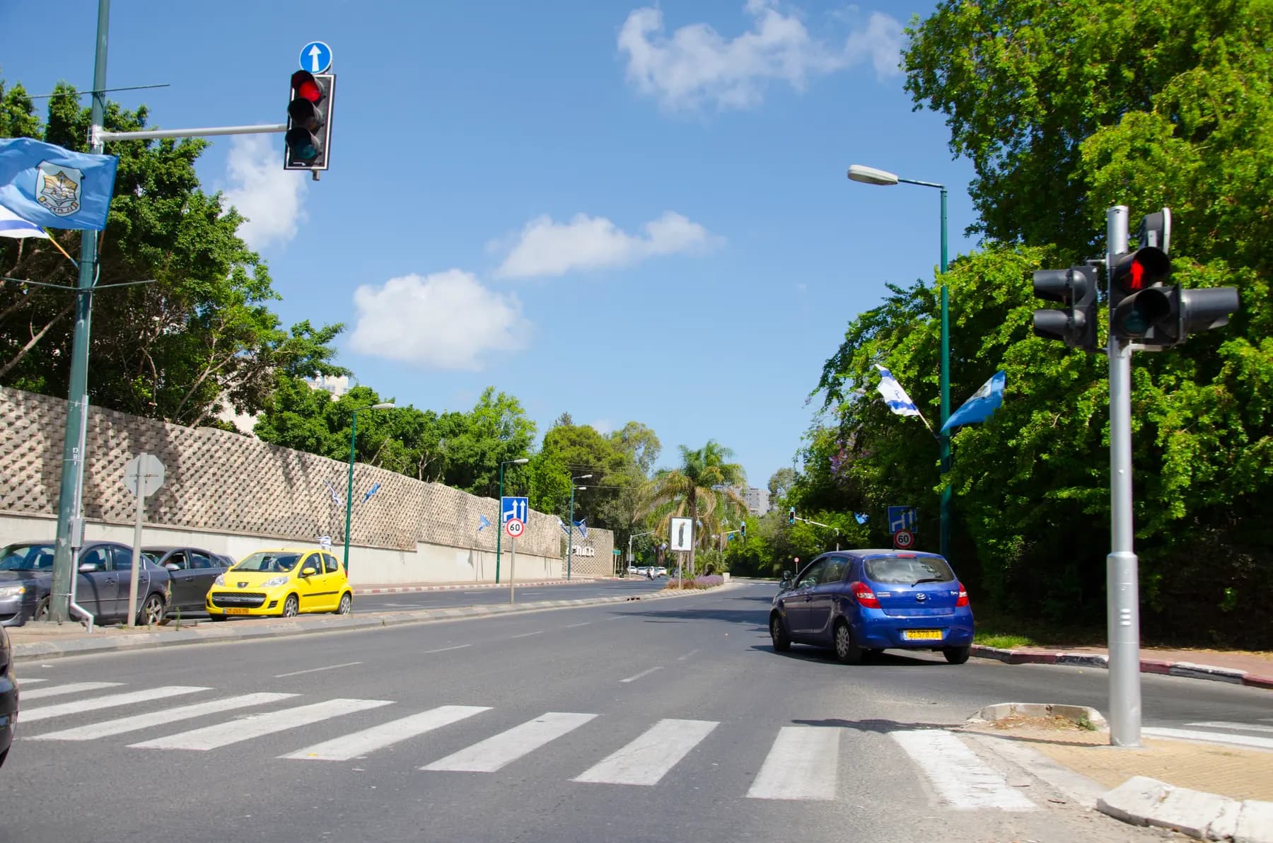 A tree-lined street in Givatayim with a pedestrian crossing, traffic lights and flags on the lamp posts.
