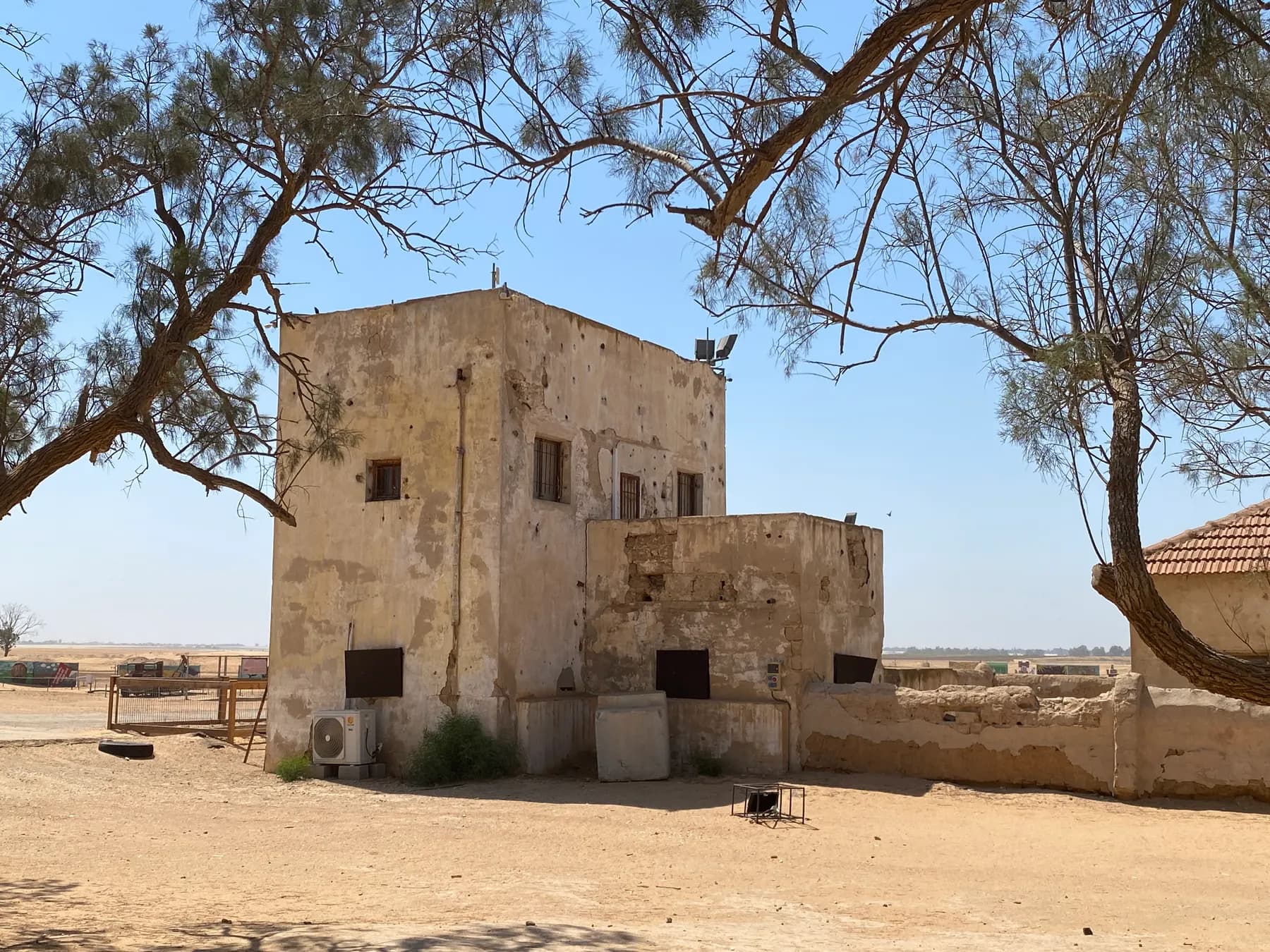 An old two-storey plastered building with a lower wing beside it in Gvulot, under the branches of a tree, with open desert ground in front.