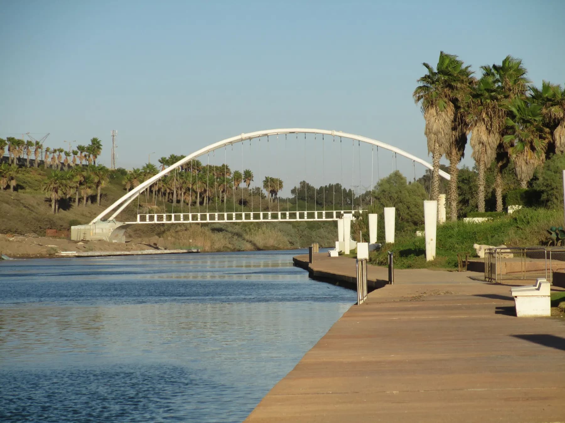 A white arched footbridge over the Hadera stream, with a riverside promenade and palm trees