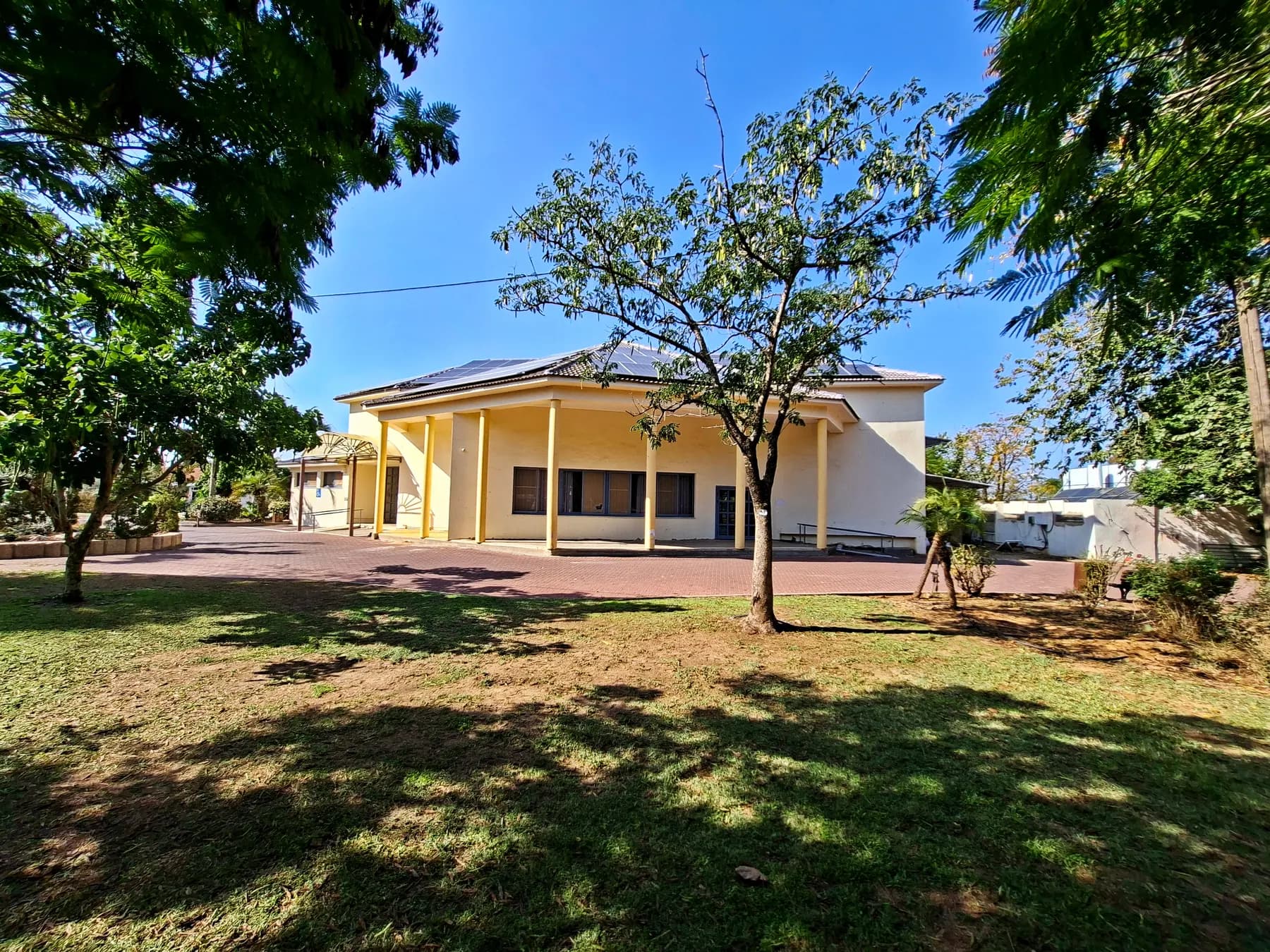 The community hall of Hagor, a pale yellow building with columns, seen across a lawn under shade trees.