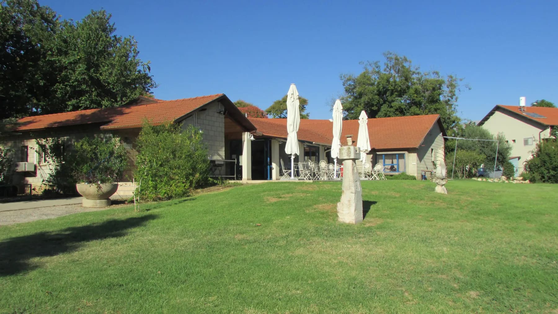 A wide lawn with stone sculptures in HaGoshrim, in front of low buildings with red tiled roofs and folded white parasols, under a blue sky.