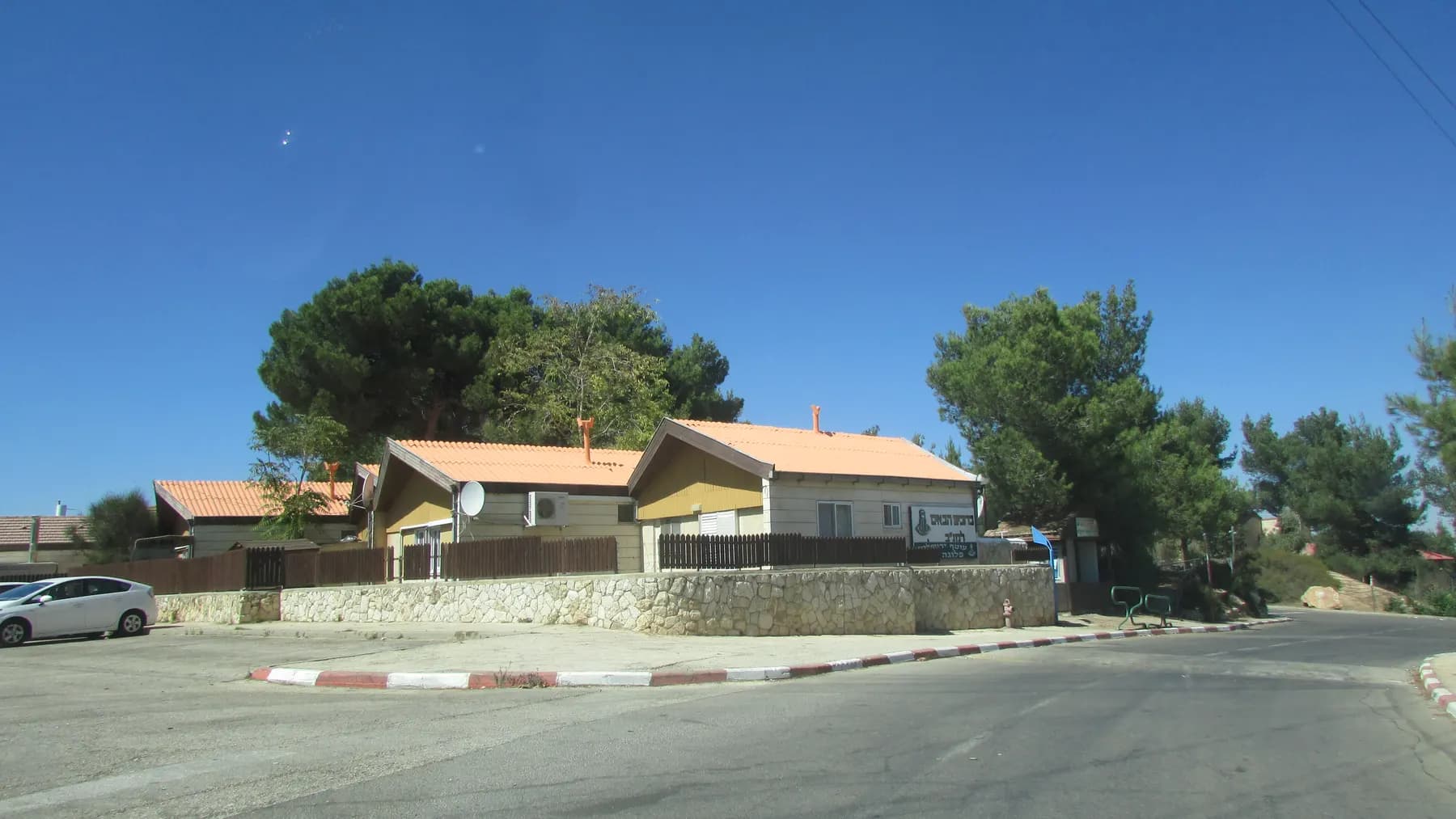 Small houses with orange tiled roofs behind a low stone wall on a street corner in Har Gilo, among pine trees.