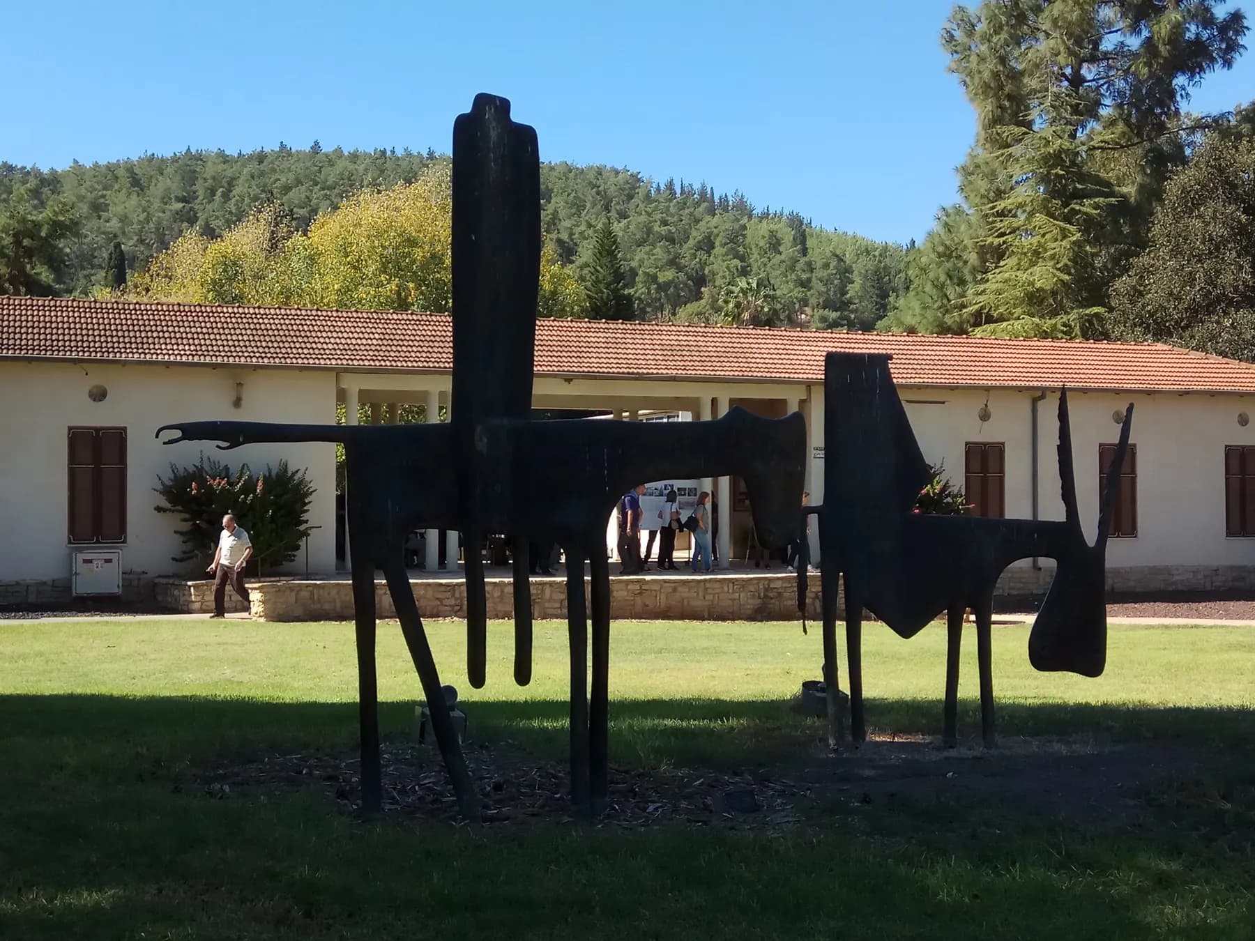 A dark metal sculpture of Don Quixote and Sancho Panza on a lawn in HaZore'a, in front of a long, low building with a tiled roof and a wooded hill.
