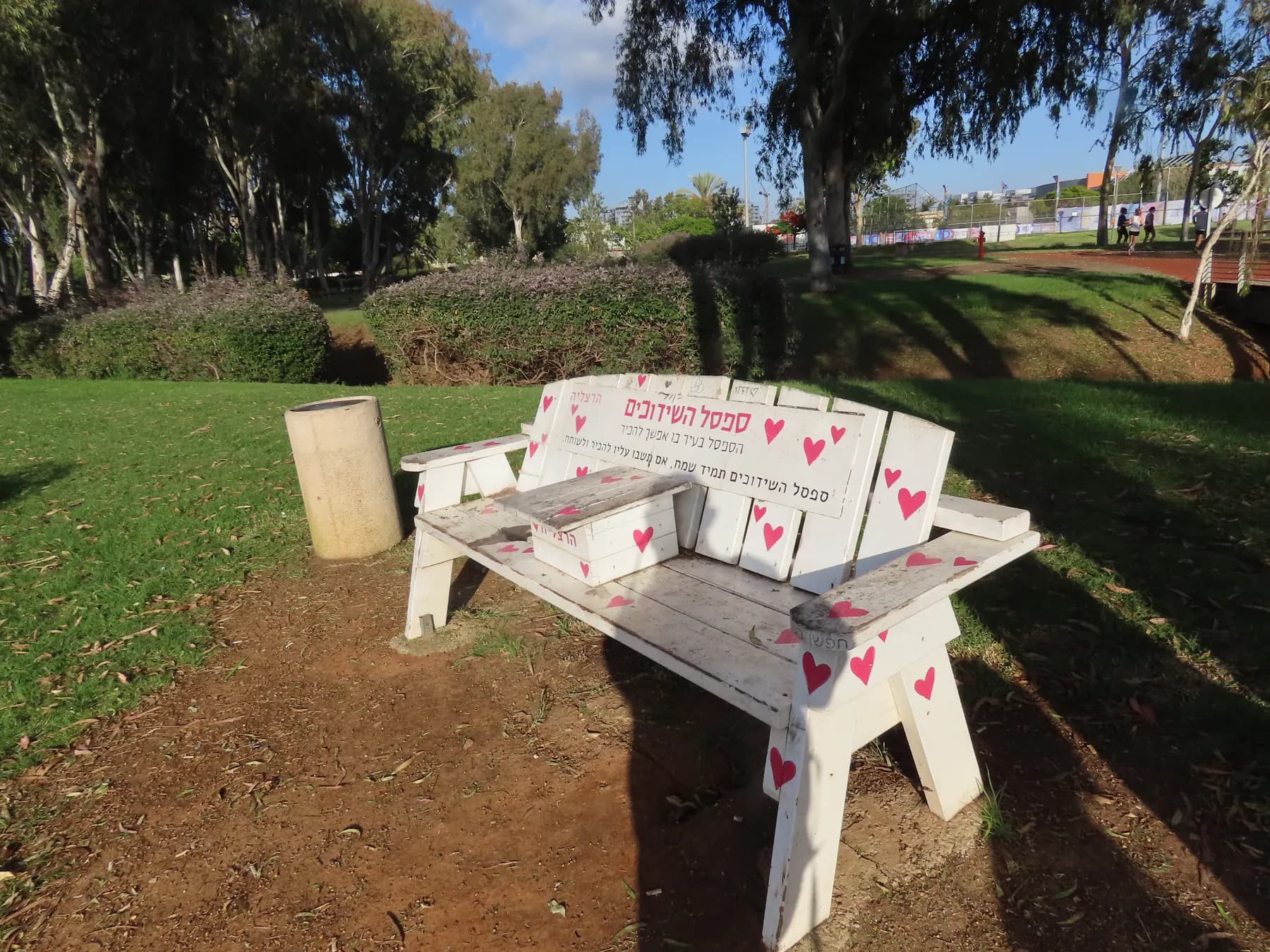 A white bench painted with hearts on the lawn of Herzliya Park, under the trees