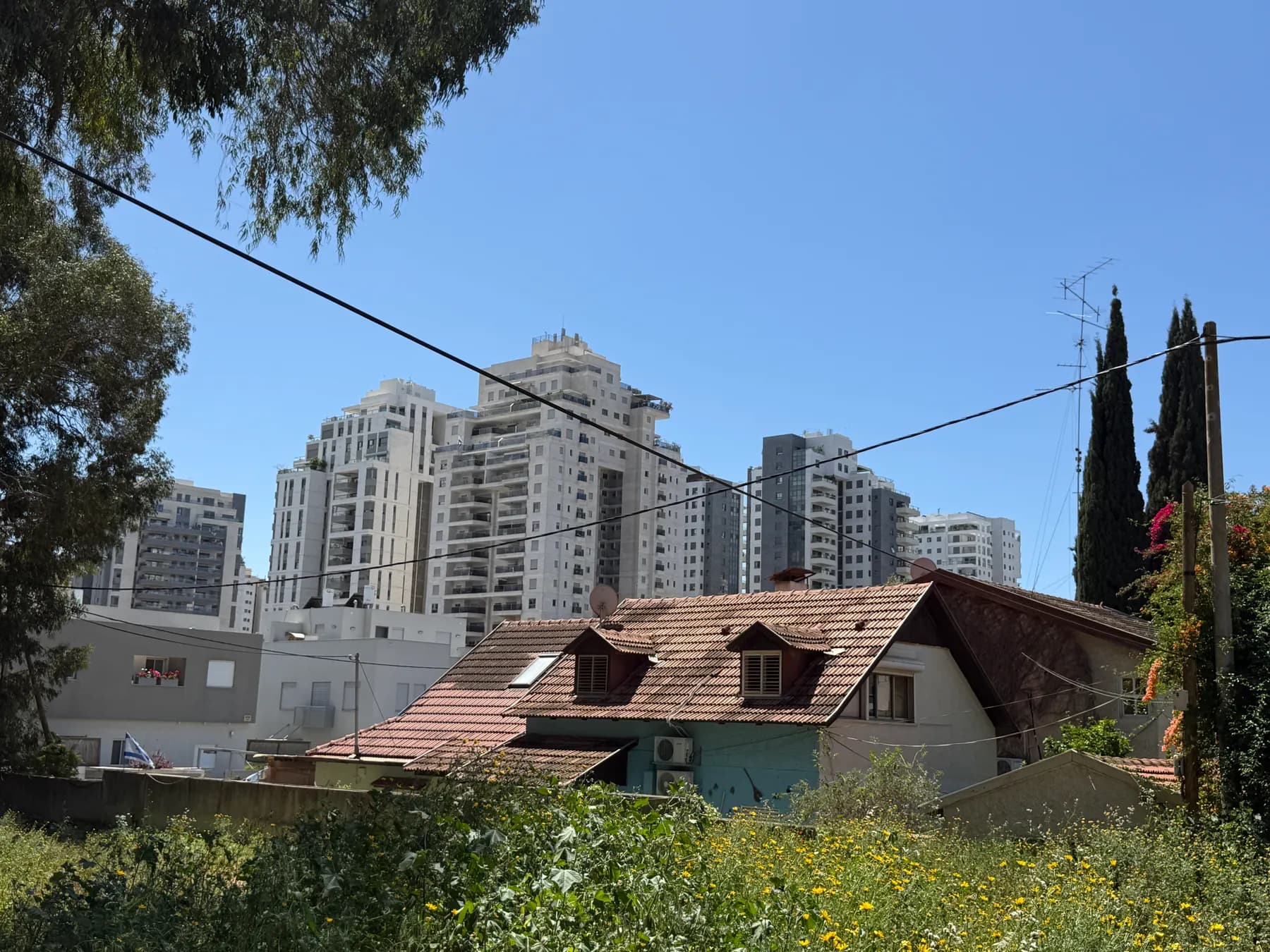 An older house with a tiled roof in Hod Hasharon, with new apartment towers rising behind it.