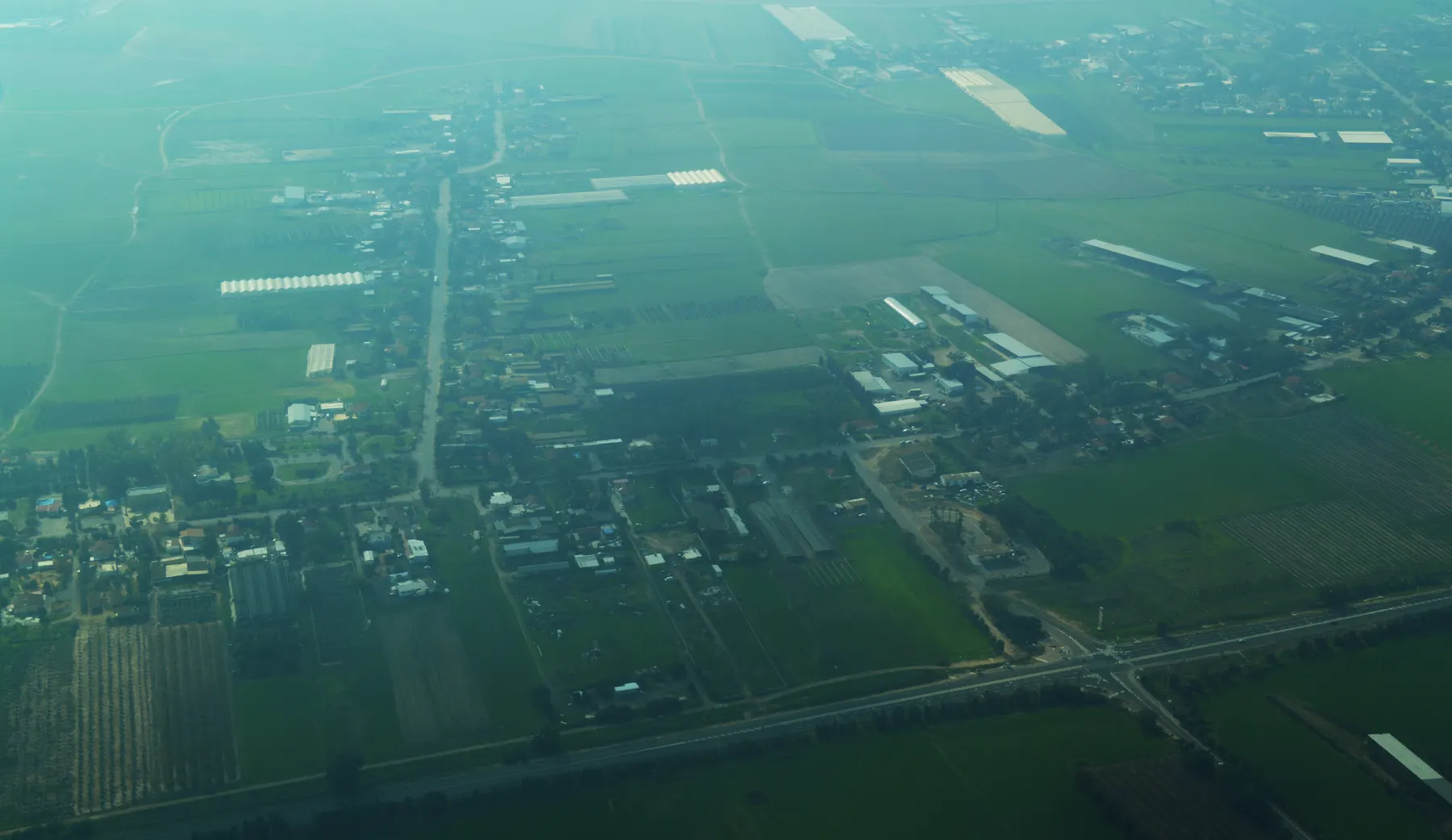 An aerial view of Hodaya on a hazy day, with houses and farm buildings along its roads, greenhouses and green fields around them, and a highway in the foreground.