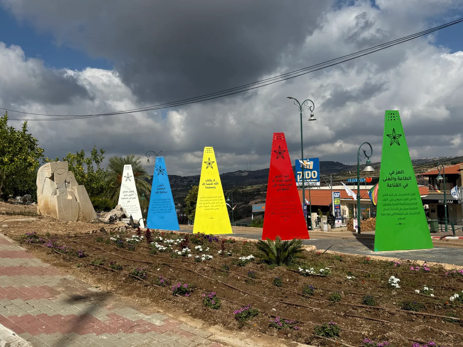 Five tall painted cones in white, blue, yellow, red and green at the entrance to Hurfeish, with a flower bed in front and shops behind.
