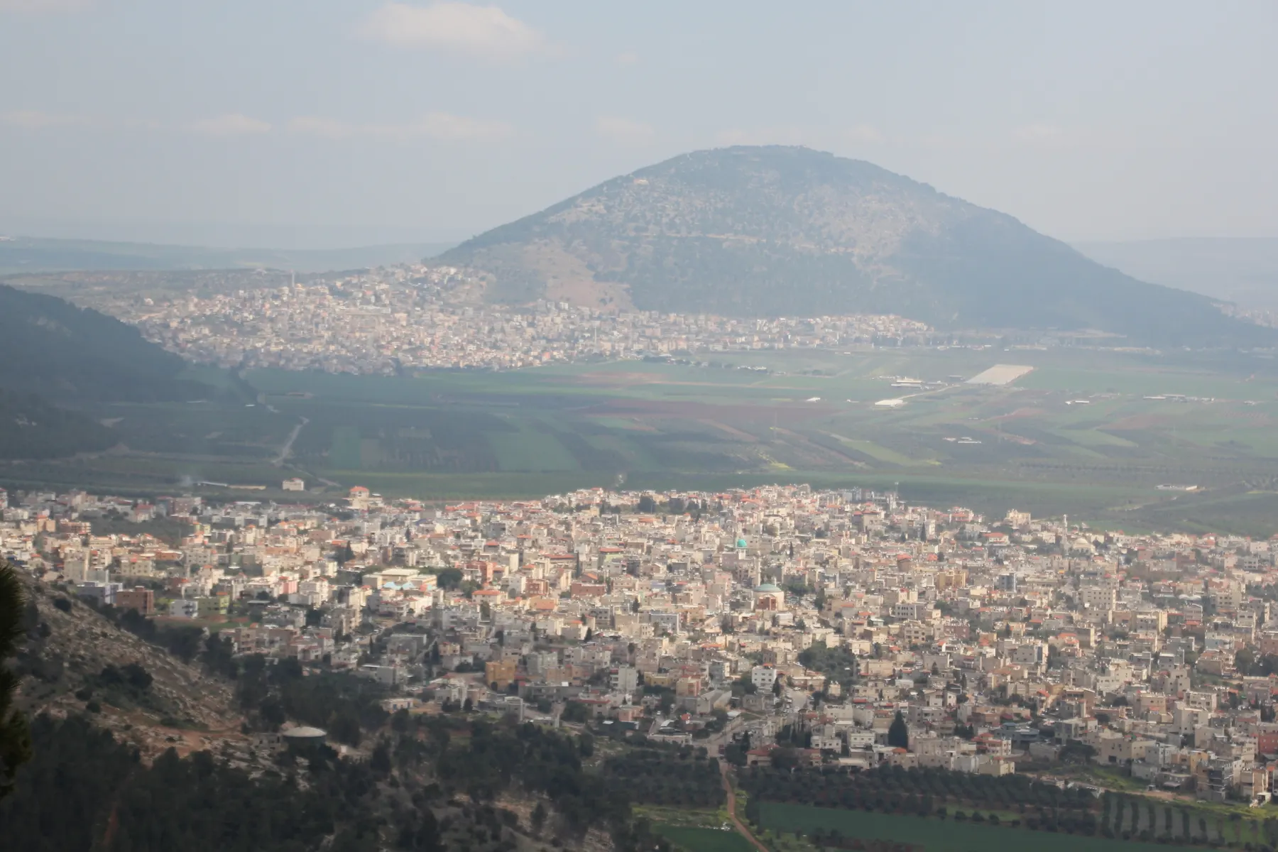 The dense houses of Iksal seen from Mount Precipice, with farm fields, another town and a rounded mountain behind.