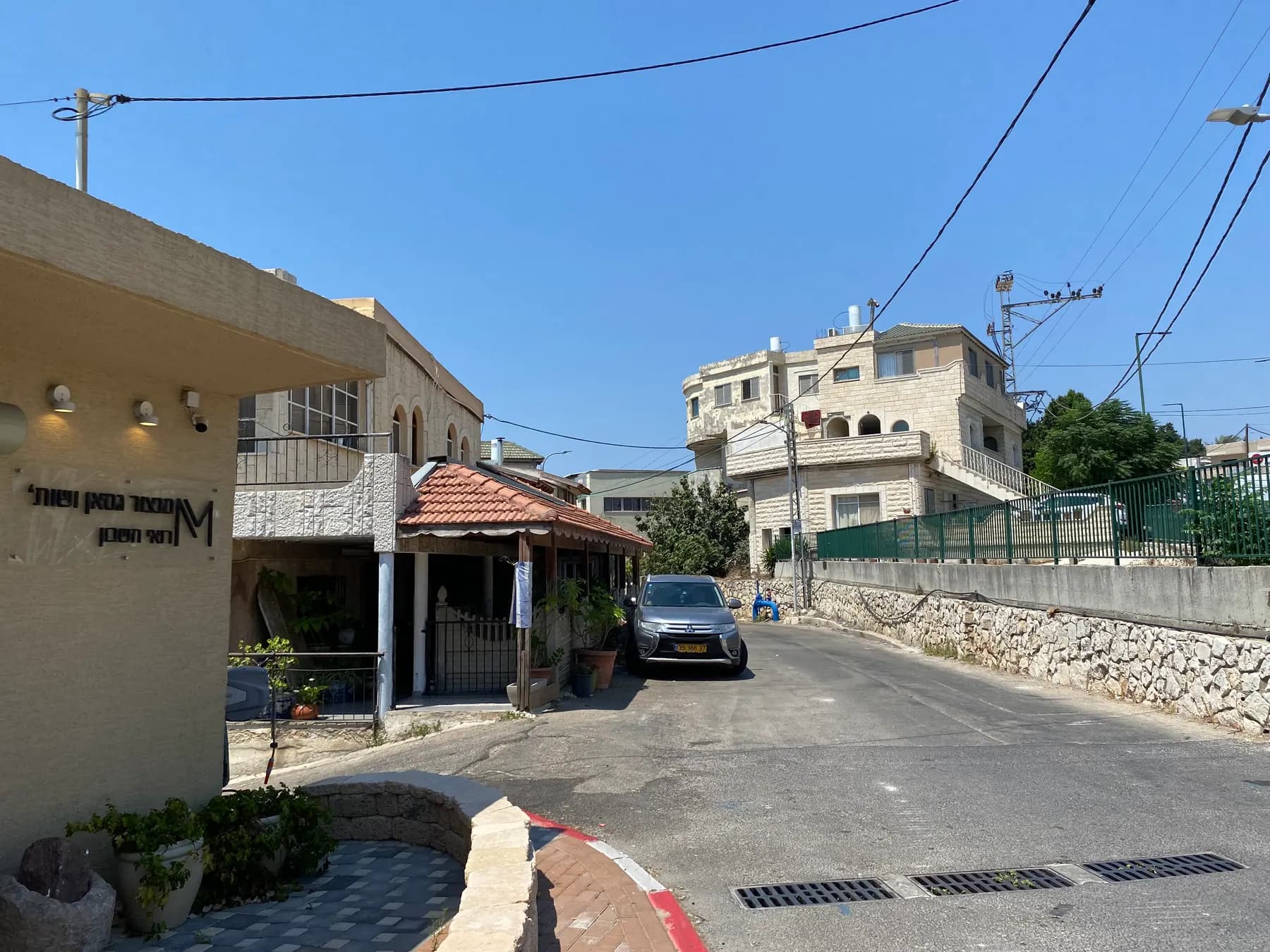 Al-Hikmah Street in Isfiya, with stone houses, a parked car and trees under a blue sky.