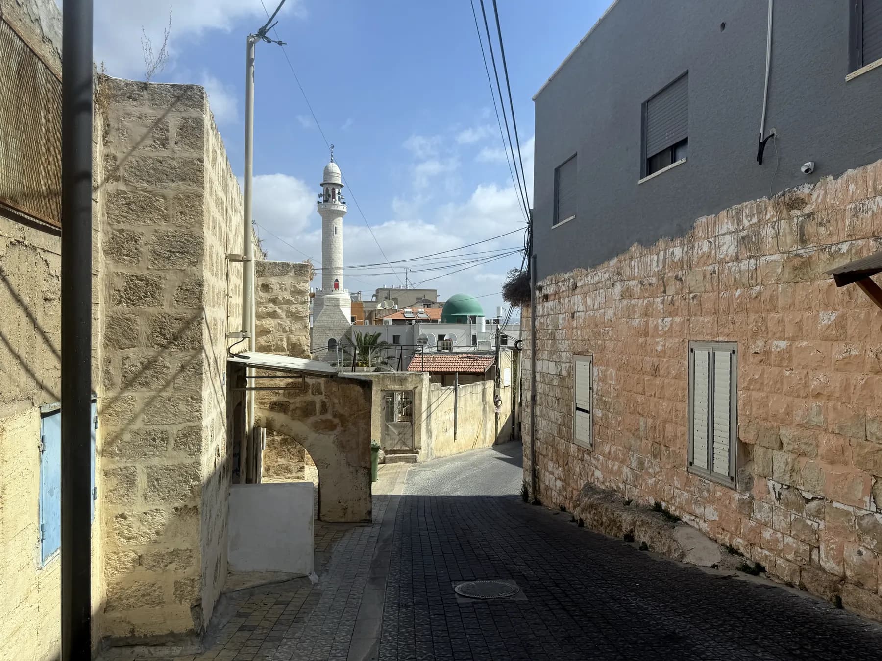 A narrow paved lane between stone houses in Jadeidi-Makr, with the minaret and green dome of the A-Nur Mosque at its end.