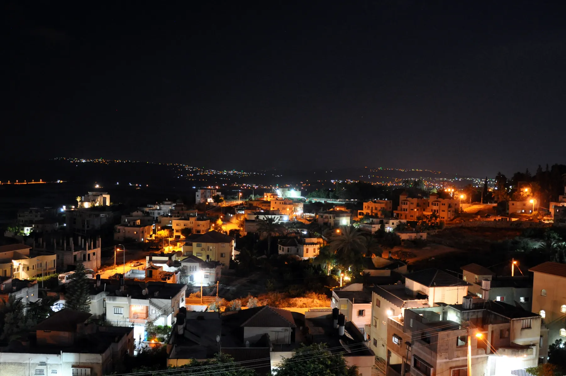 Houses on a hillside in Jatt at night, lit by street lamps, with the lights of other towns on the hills beyond.