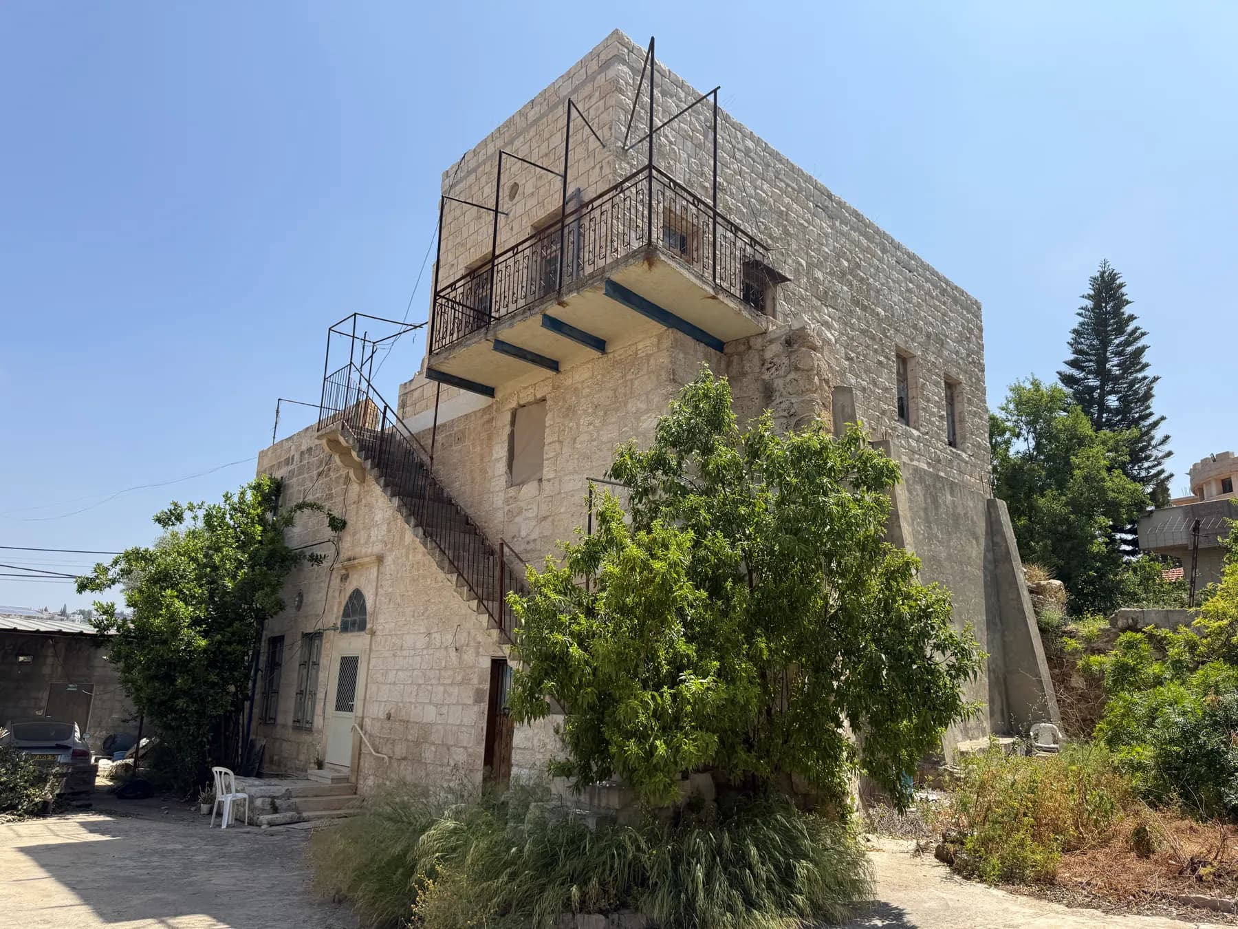 An old two-storey stone house with an outside staircase and a balcony, among trees and shrubs in Julis.
