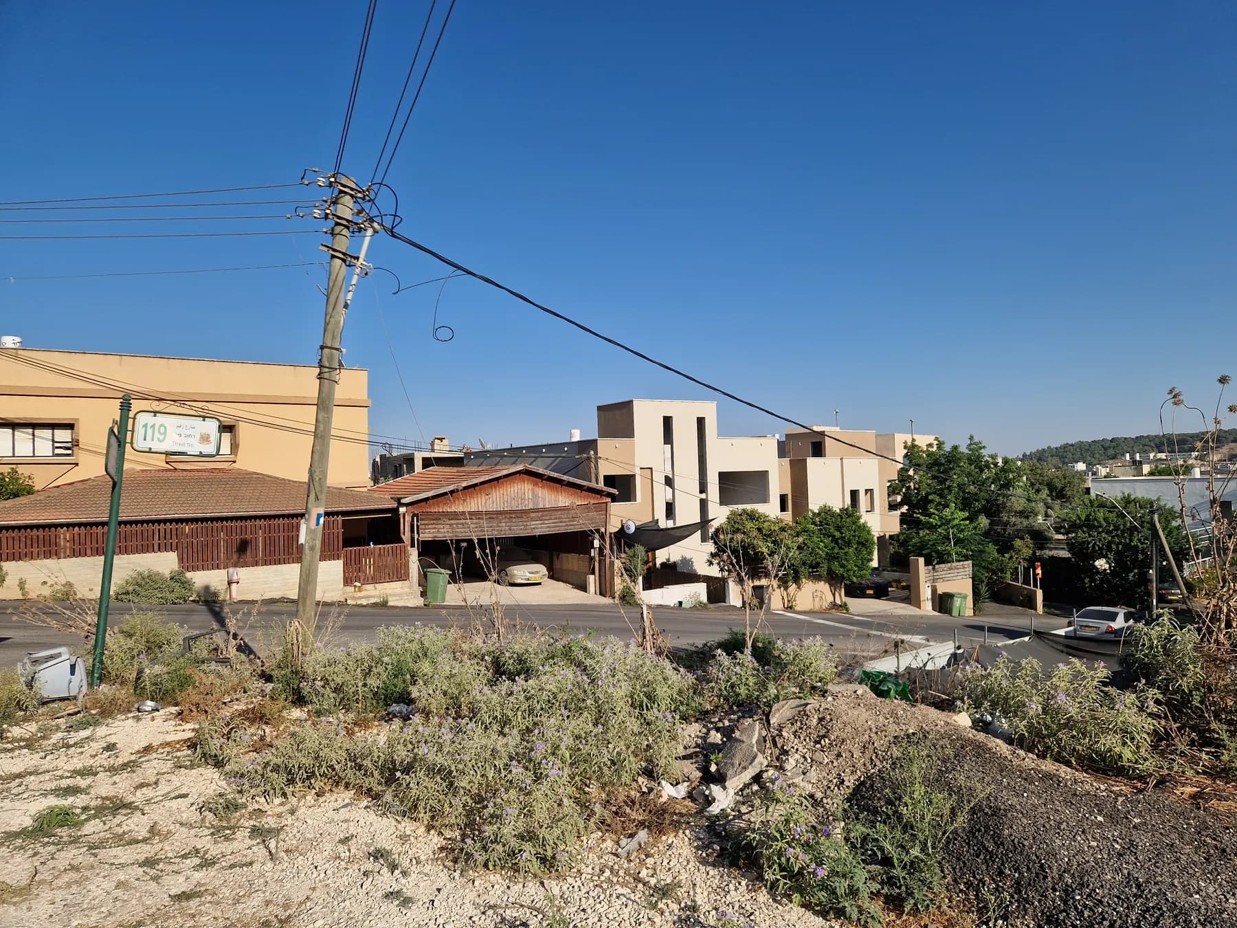 A street in Ka'abiyye-Tabbash-Hajajre lined with two- and three-storey houses, with a utility pole and dry scrub in the foreground.