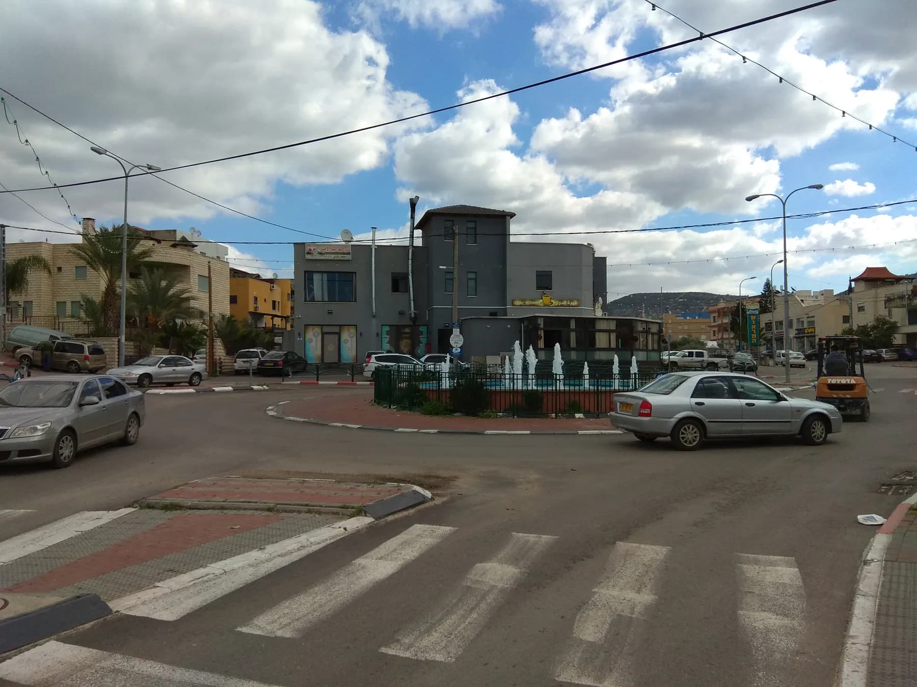 A roundabout with a fountain in Kabul, with shops, houses and parked cars around it under a partly cloudy sky.