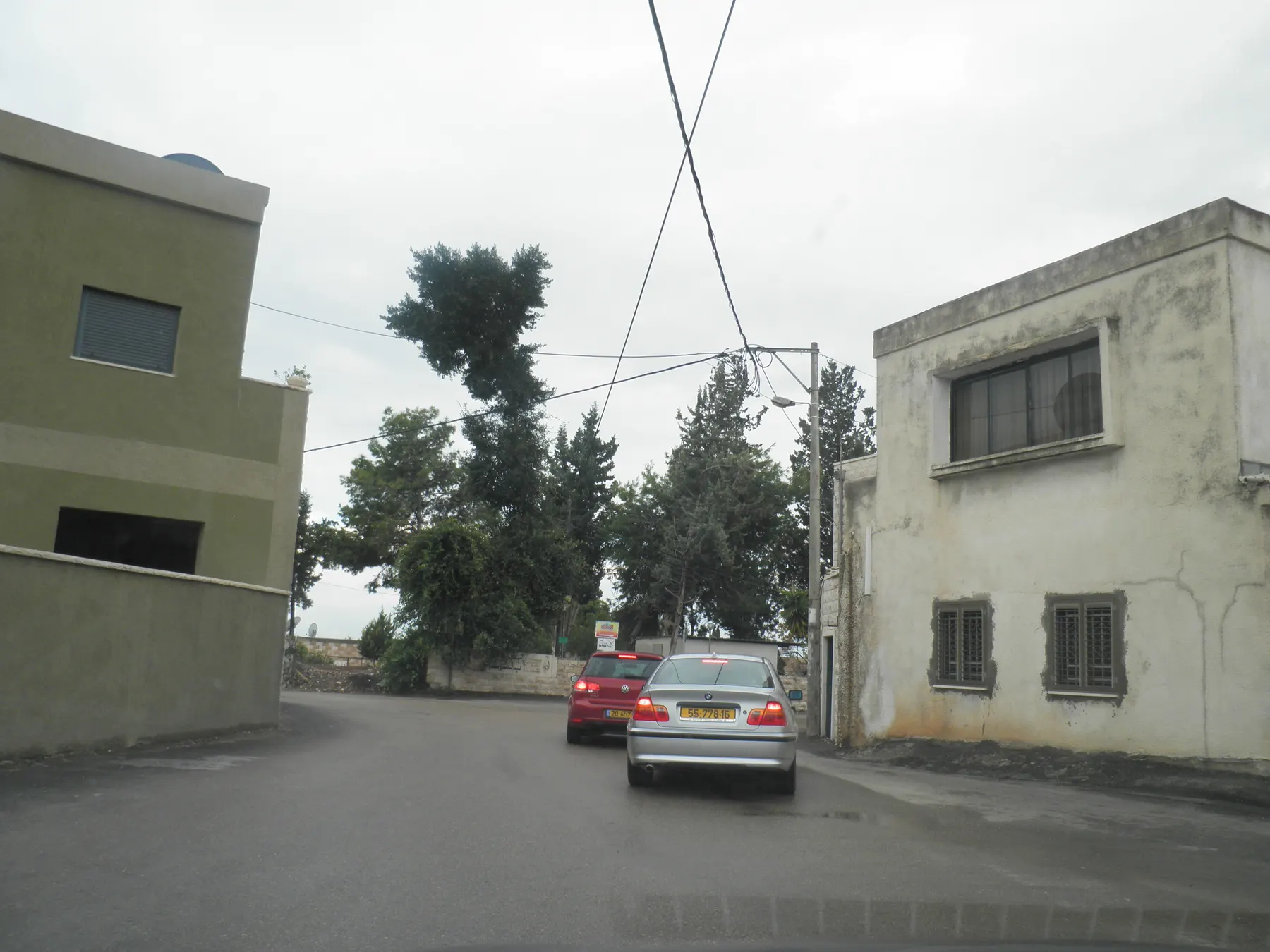 Two cars on a narrow street in Kafr Qara between two-storey houses, with tall trees ahead on an overcast day.