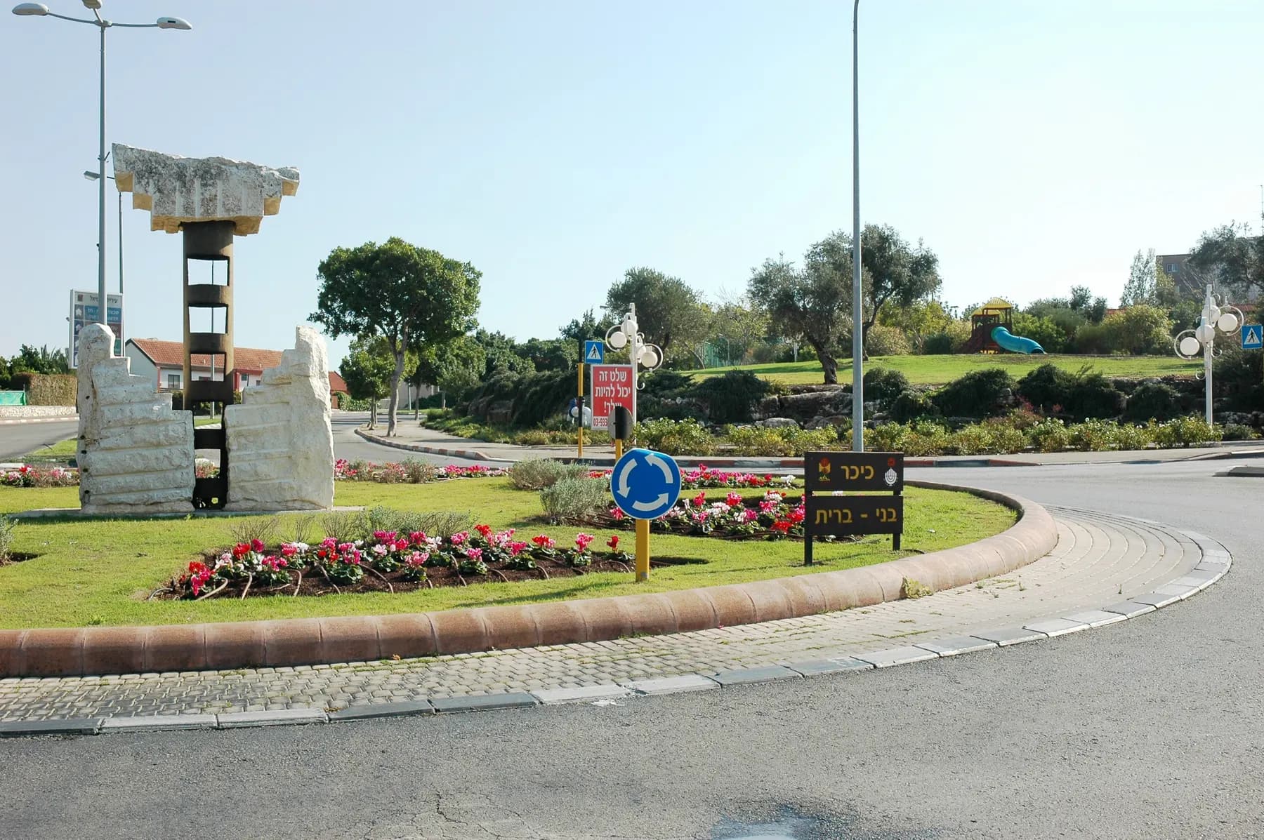 Bnei Brit Square in Karmiel, a roundabout with a stone sculpture, flower beds and olive trees.