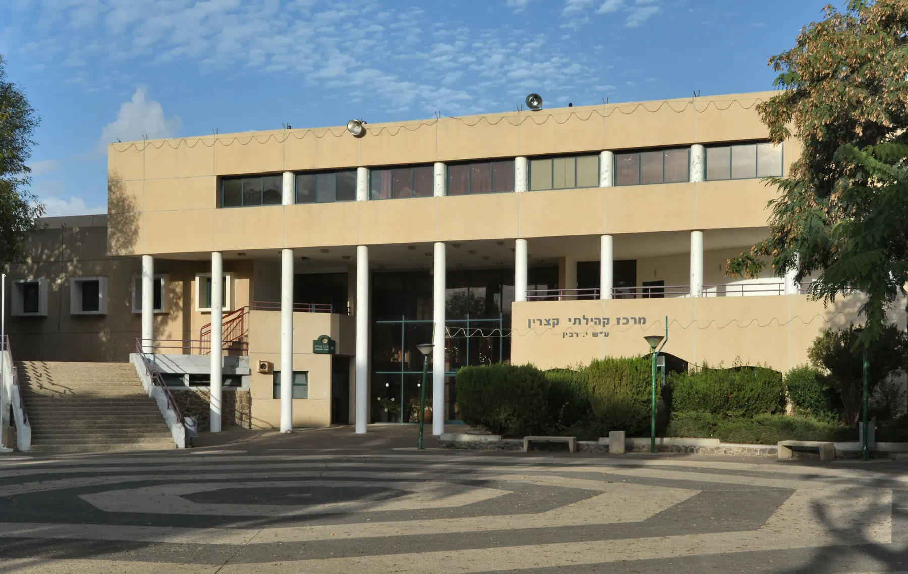 The community centre of Katzrin, a pale building with tall white columns facing a paved square with a swirling pattern.