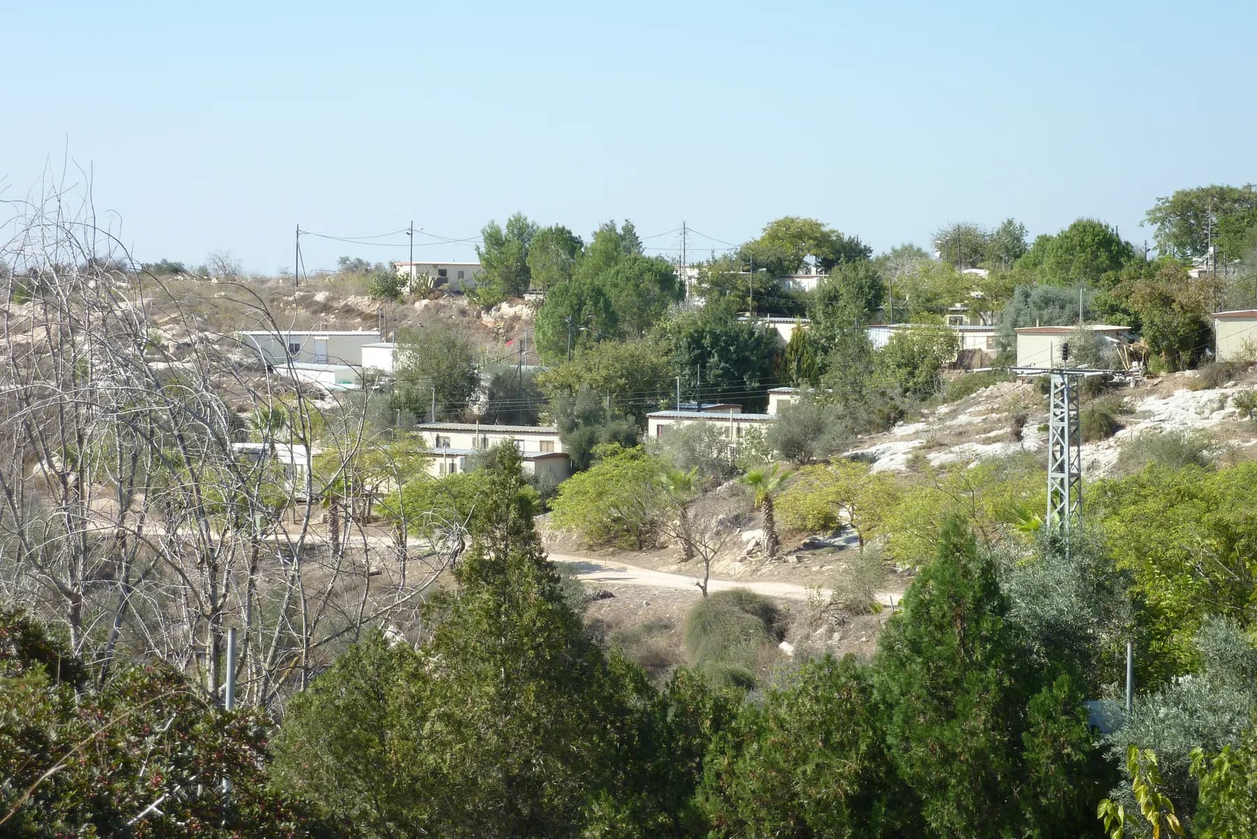 Single-storey prefabricated houses among green trees on a rocky hillside in Kedumim, with a dirt track and a power pylon below them.