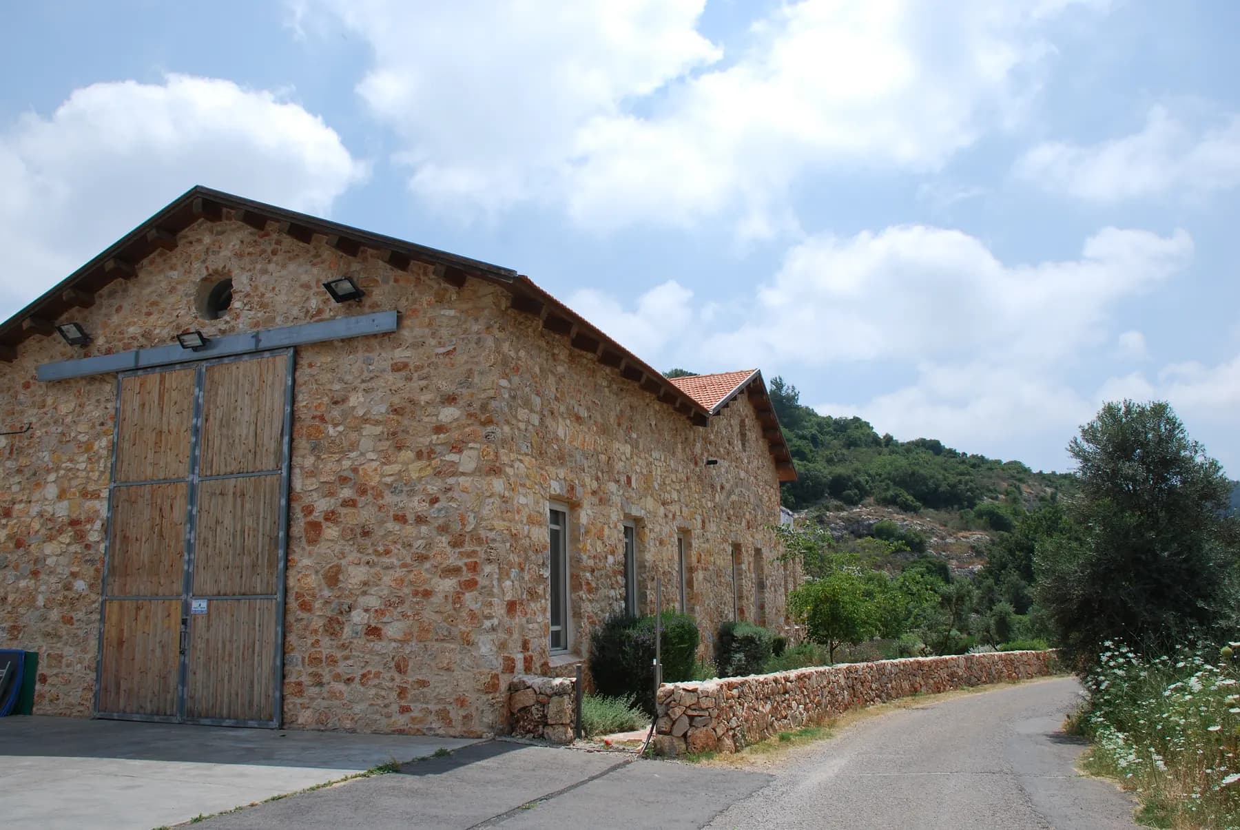 A long old stone building with a tiled roof and tall wooden doors beside a quiet road in Kerem Maharal, with green wooded hills behind.