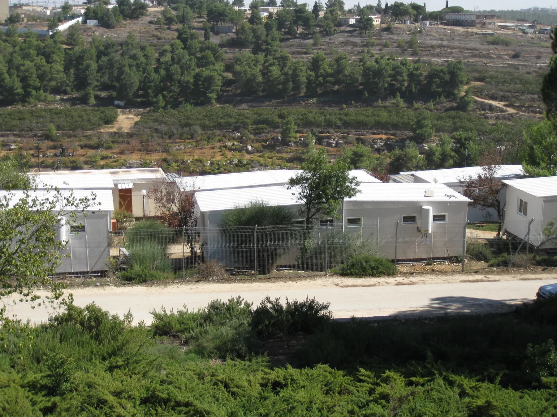 White prefabricated buildings beside a road in Kfar Etzion, with a terraced hillside of trees behind them.