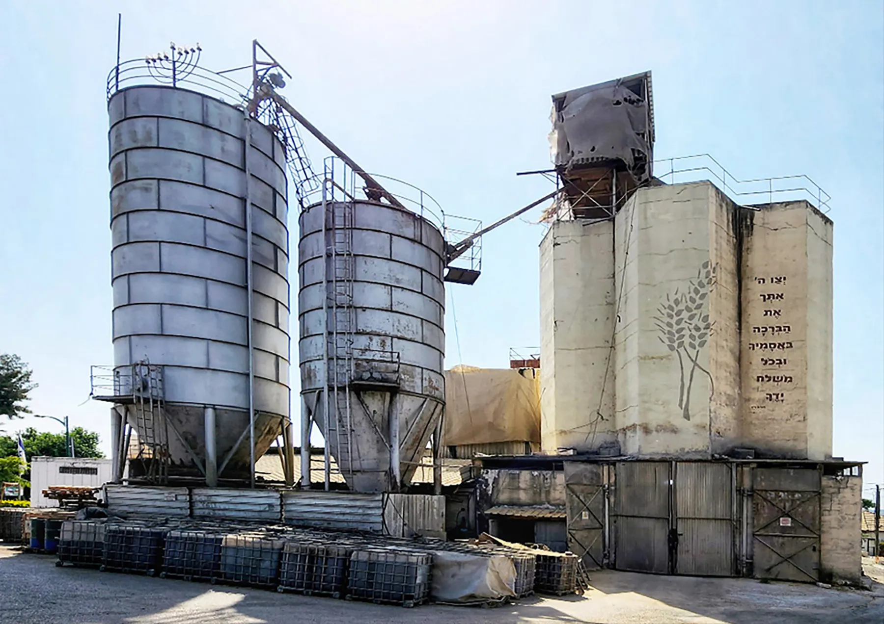 Tall metal and concrete silos beside a yard of stacked containers in Kfar Haroeh, under a blue sky.