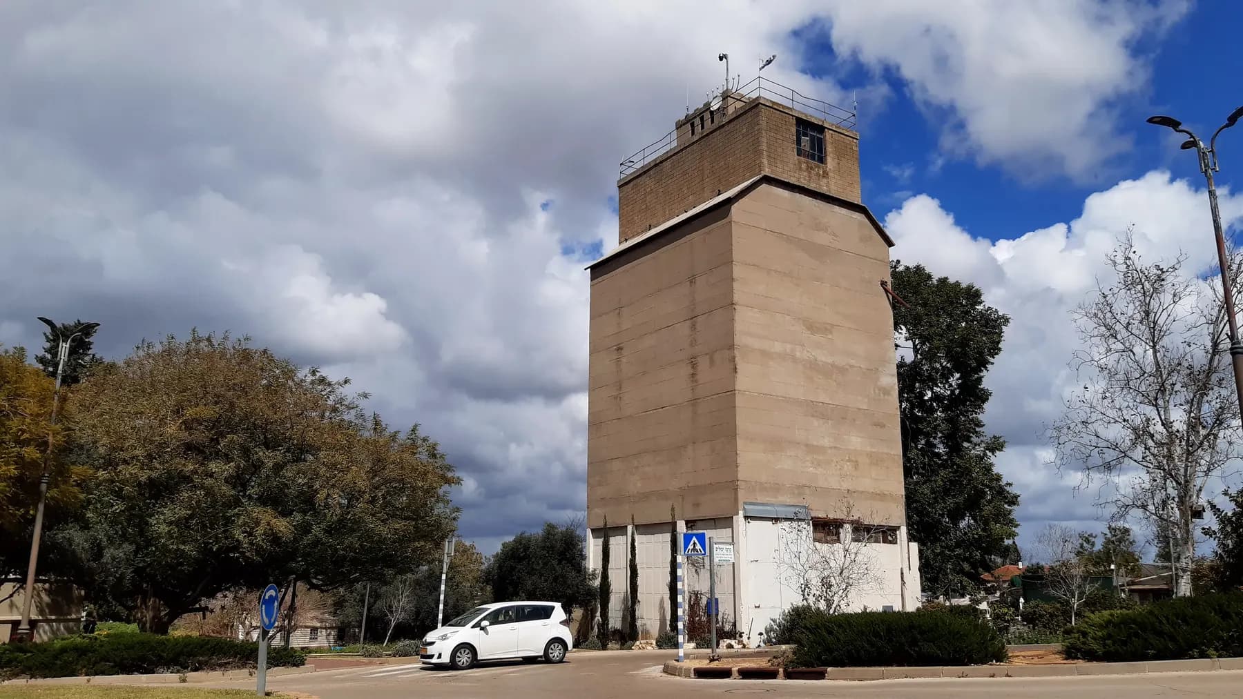 The tall concrete silo tower of Kfar Hess beside a road with a pedestrian crossing, with trees and a white car in front under a cloudy sky.