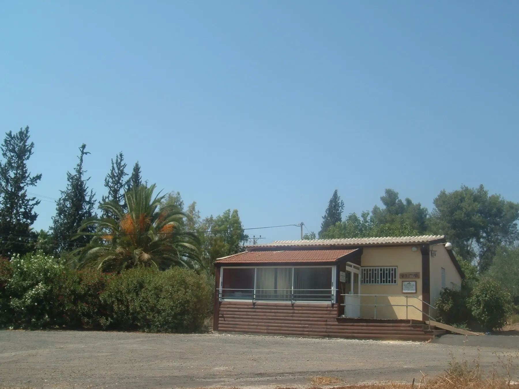 The founders' house in Kfar Kisch, a small single-storey building with a glazed porch, among palms, cypresses and pines beside a gravel yard.