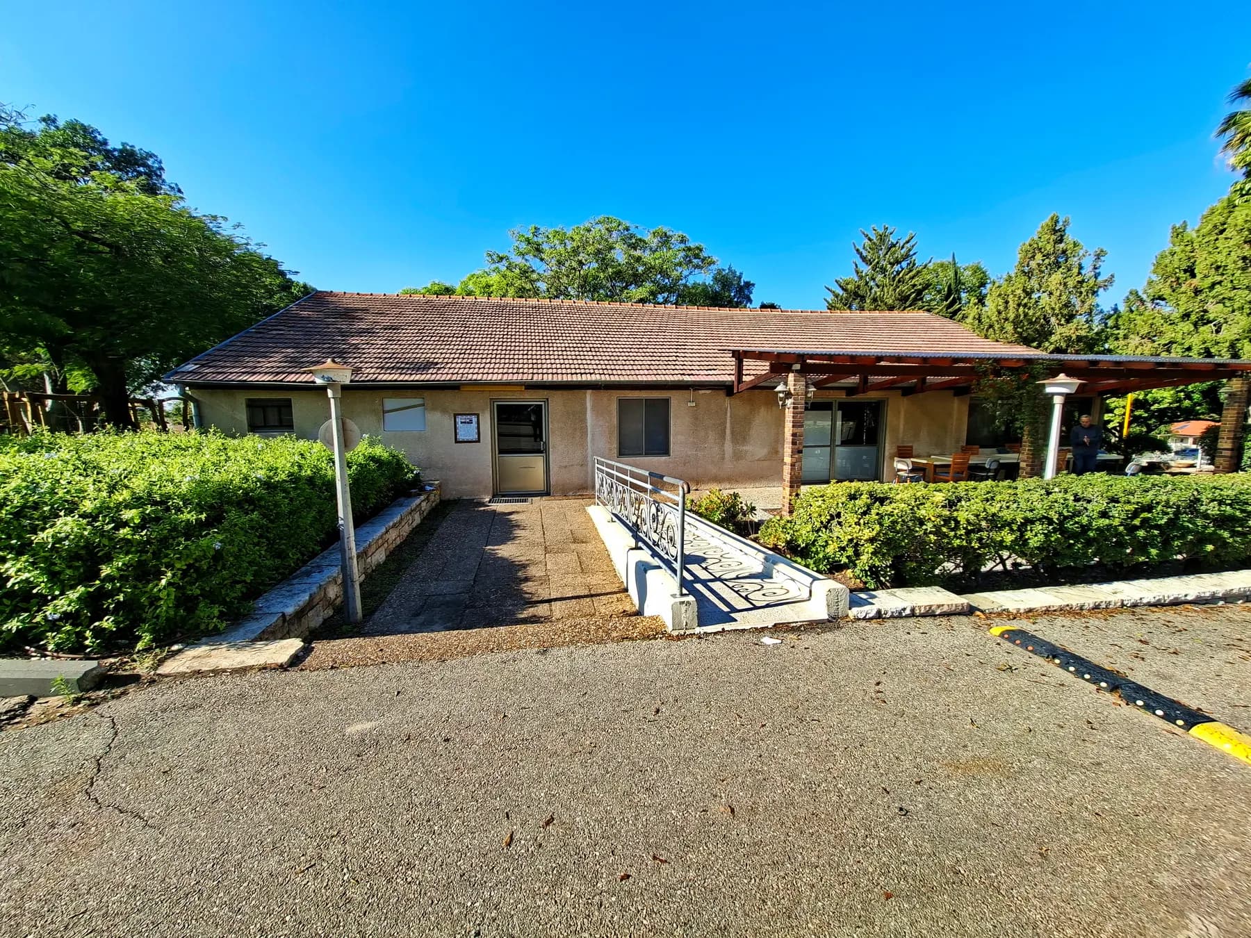 A long, low building with a tiled roof and a ramp to its door in Kfar Menahem, among hedges and tall trees.