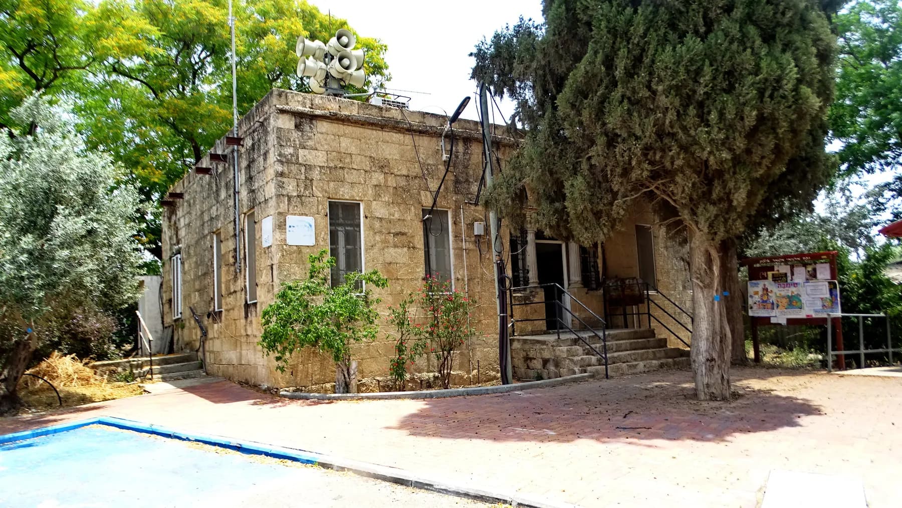 An old stone building with steps up to its entrance, shaded by tall trees, in Kfar Shmuel.