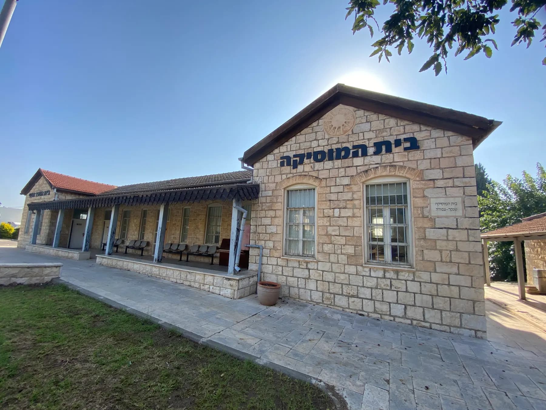 A long one-storey stone building with a tiled roof, arched windows and a covered porch, signed as the House of Music, on a lawn in Kfar Tavor.