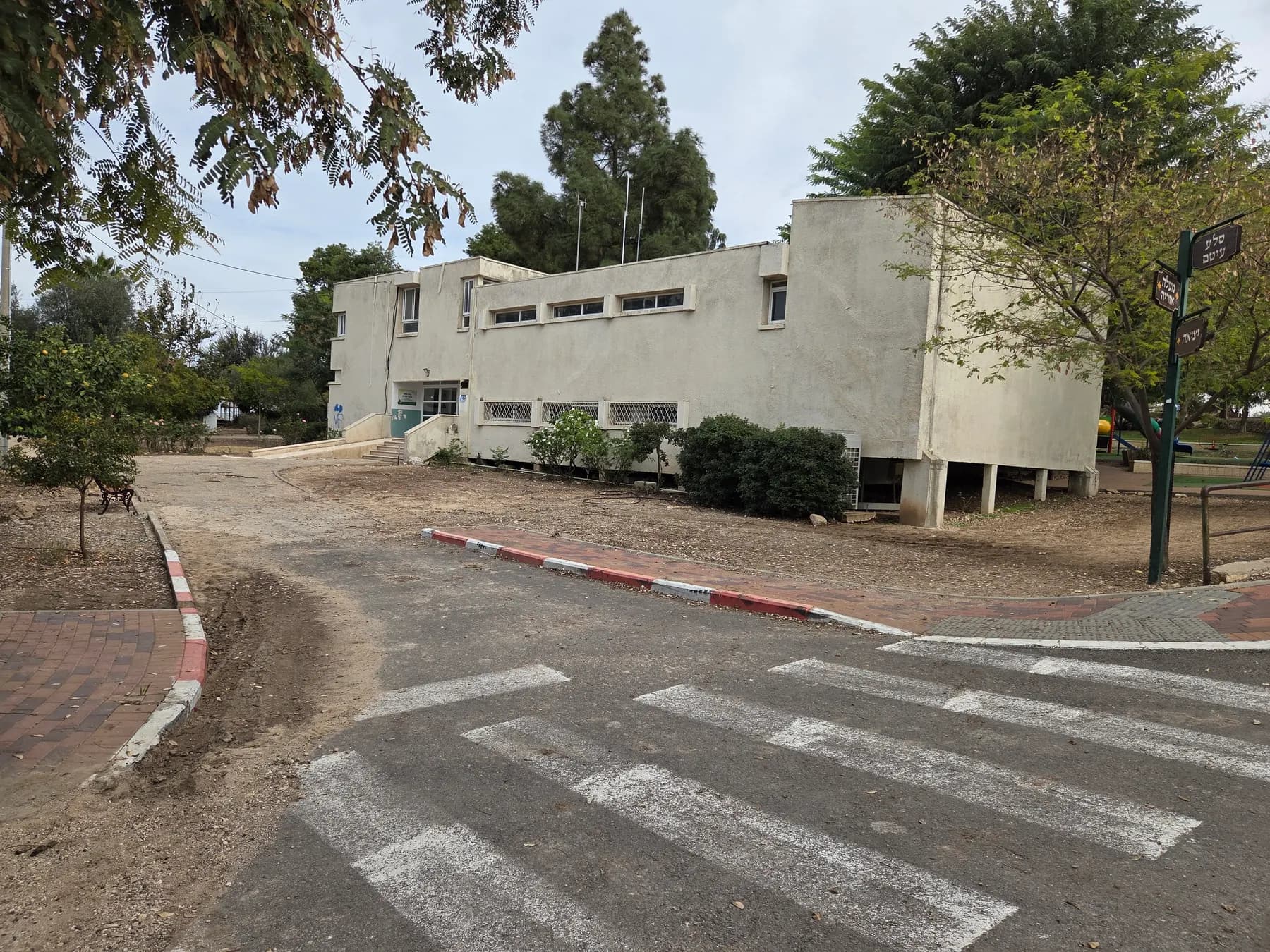A white two-storey hall partly raised on pillars in Kfar Uria, among trees beside a road with a pedestrian crossing.
