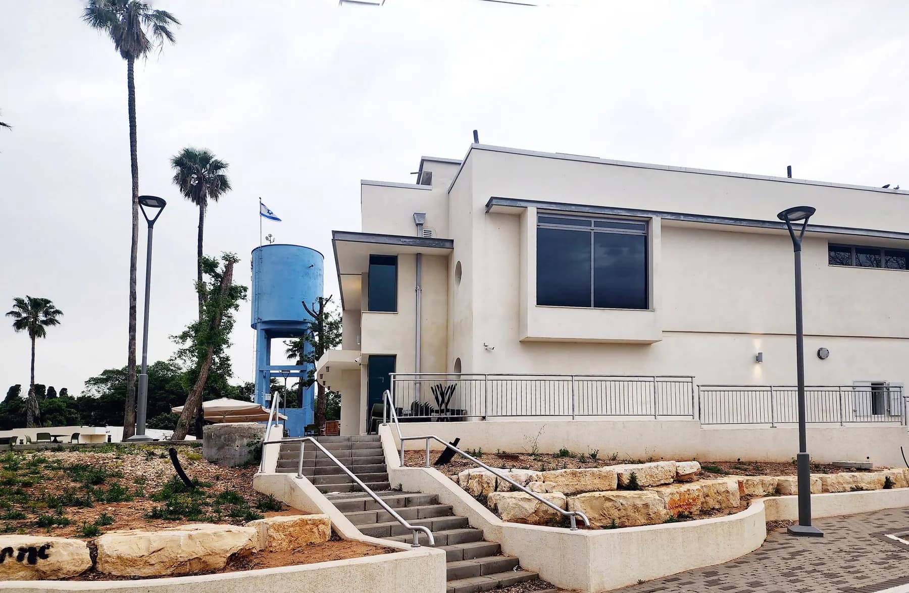 A modern white building with wide steps and a ramp in Kfar Vitkin, with palm trees and a blue water tower behind it.